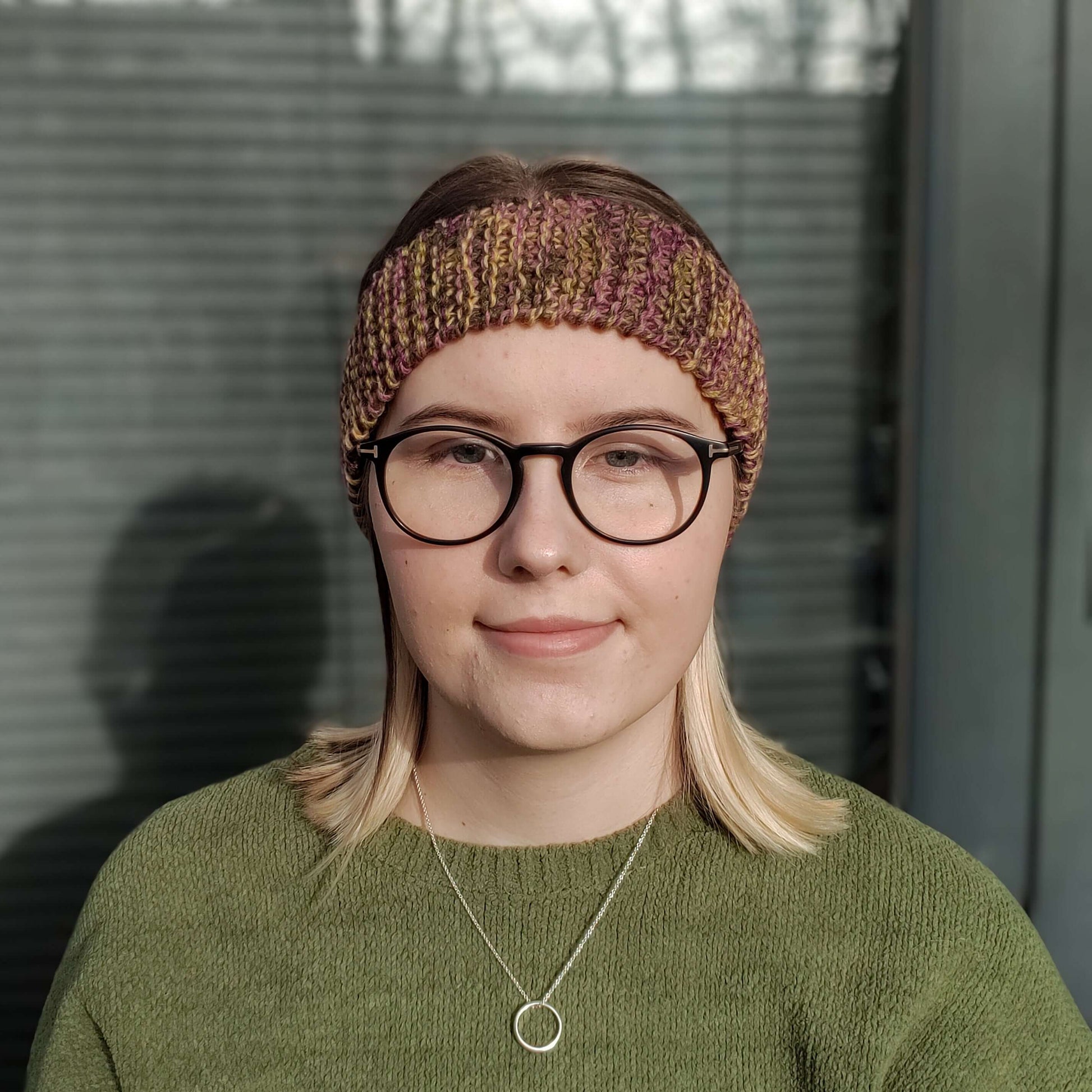 A blonde woman in a green jumper and glasses models a dark yellow green and purple variegated knitted headband