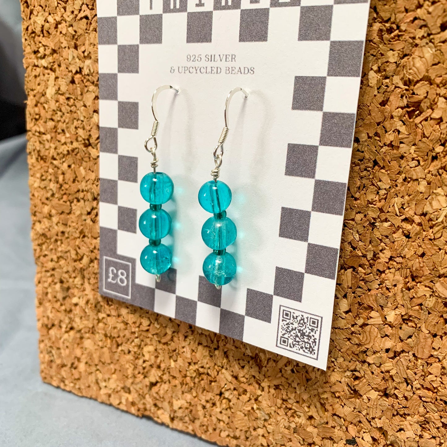 blue bead earrings on a checkerboard backing pegged to a corkboard against a blue background
