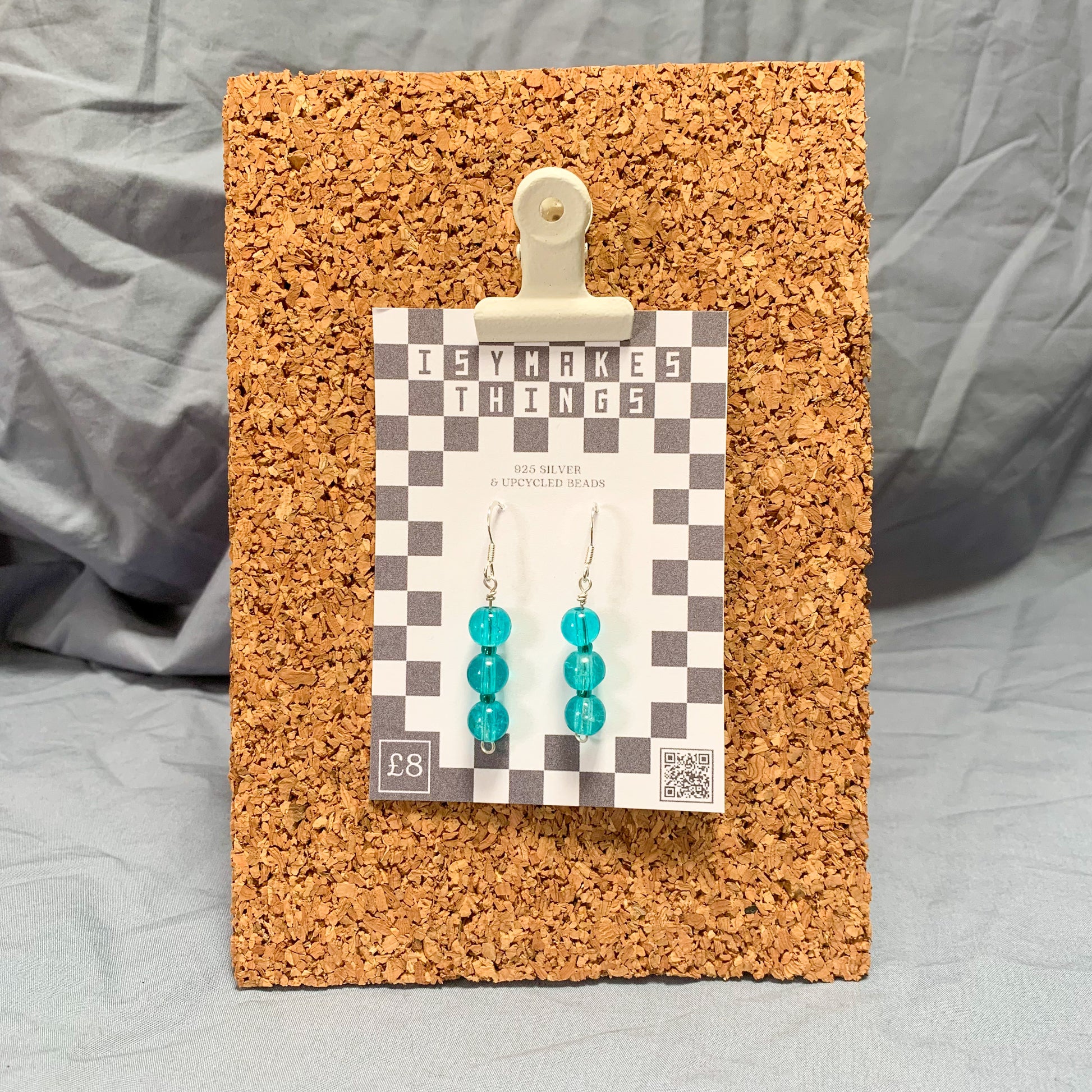 blue bead earrings on a checkerboard backing pegged to a corkboard against a blue background