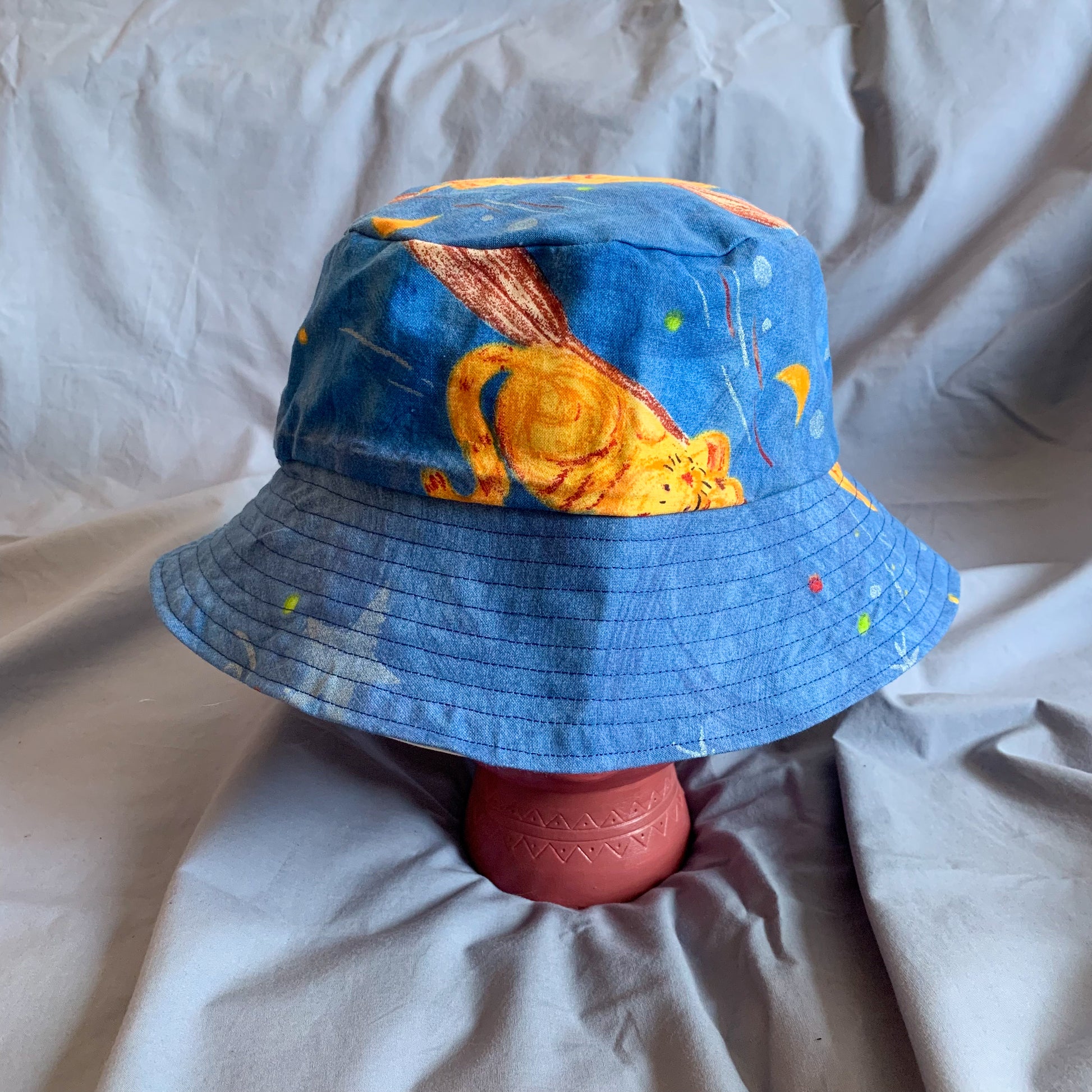 A blue bucket hat with part of a cat cartoon styled on a pink stone head against a blue background