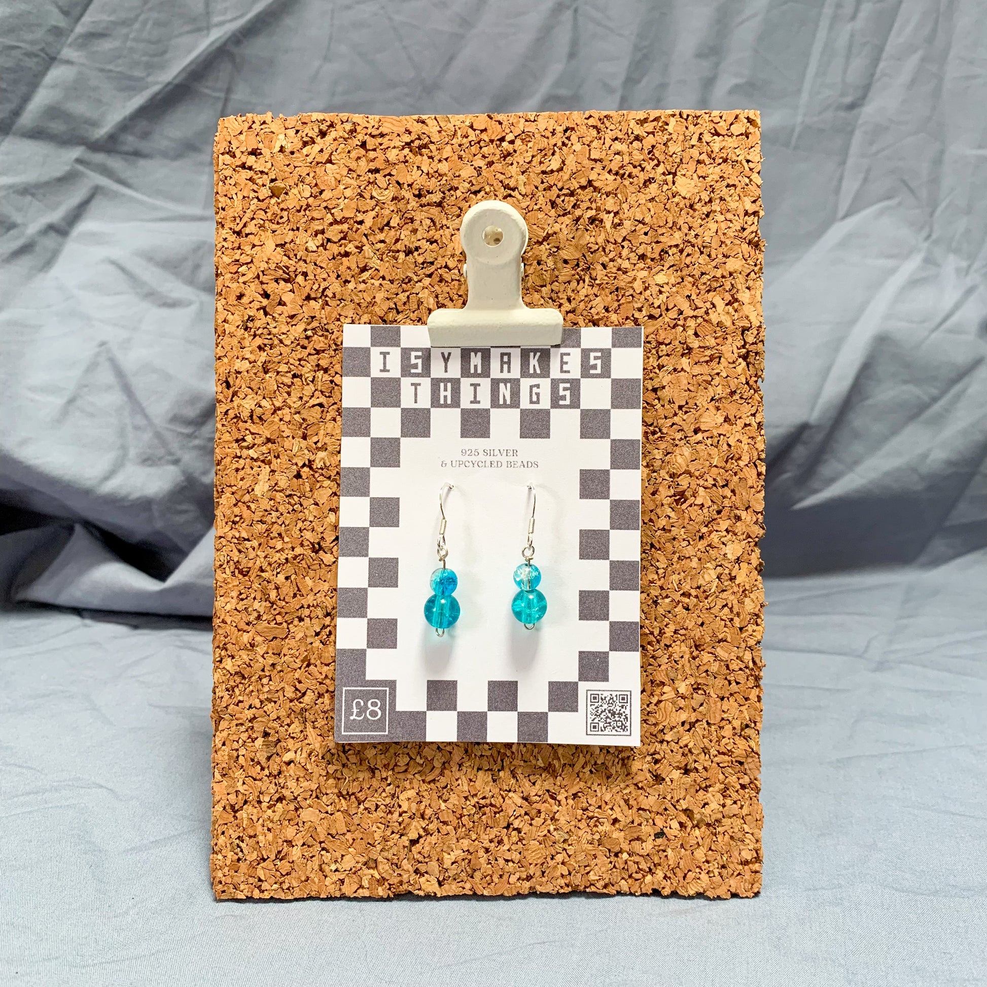 blue bead earrings on a checkerboard backing pegged to a corkboard against a blue background