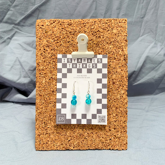 blue bead earrings on a checkerboard backing pegged to a corkboard against a blue background