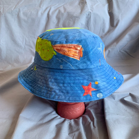 A blue bucket hat with part of a frog cartoon styled on a pink stone head against a blue background