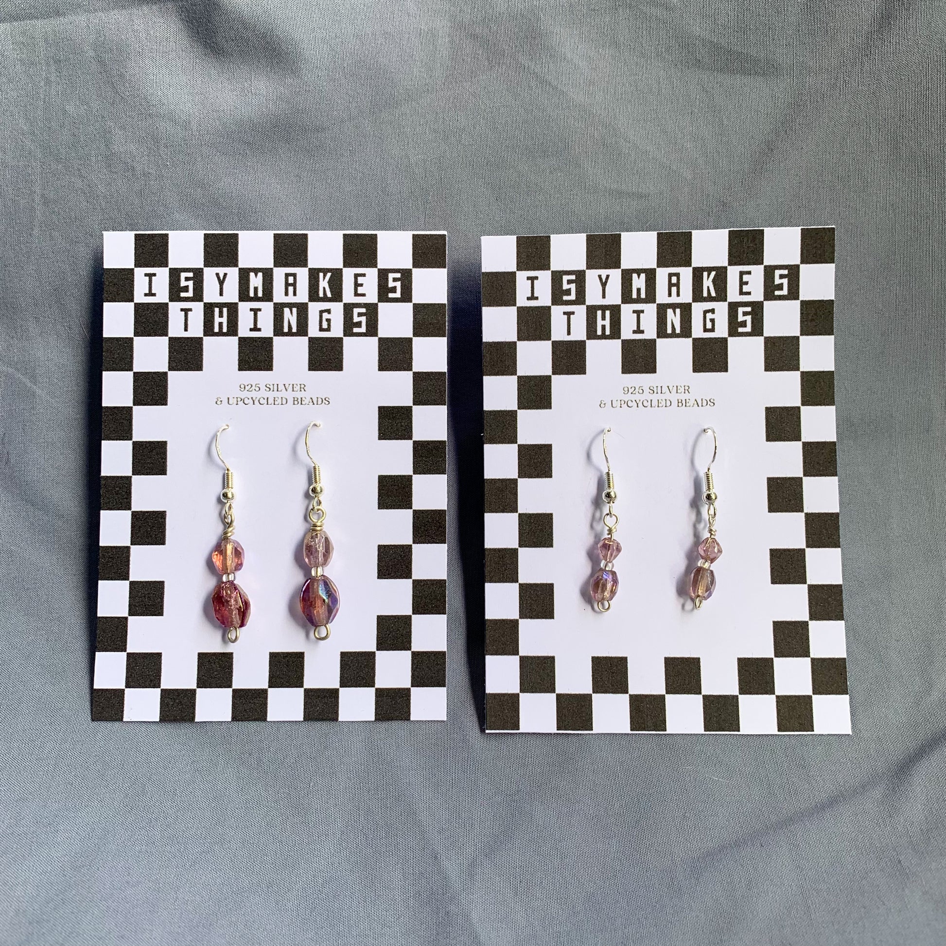 Two pairs of purple gem earrings on checkerboard backing against a blue background, both the same style but the right pair are larger and the left are smaller