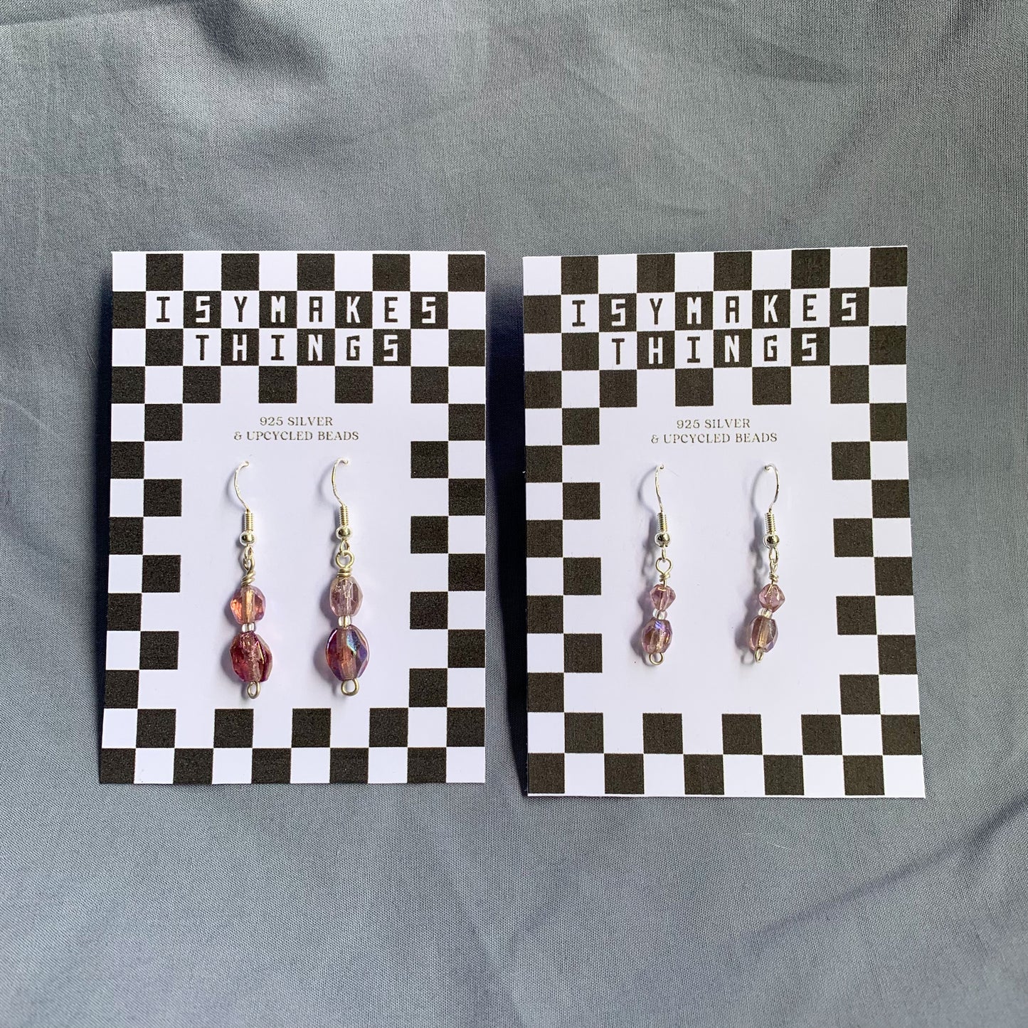 Two pairs of purple gem earrings on checkerboard backing against a blue background, both the same style but the right pair are larger and the left are smaller