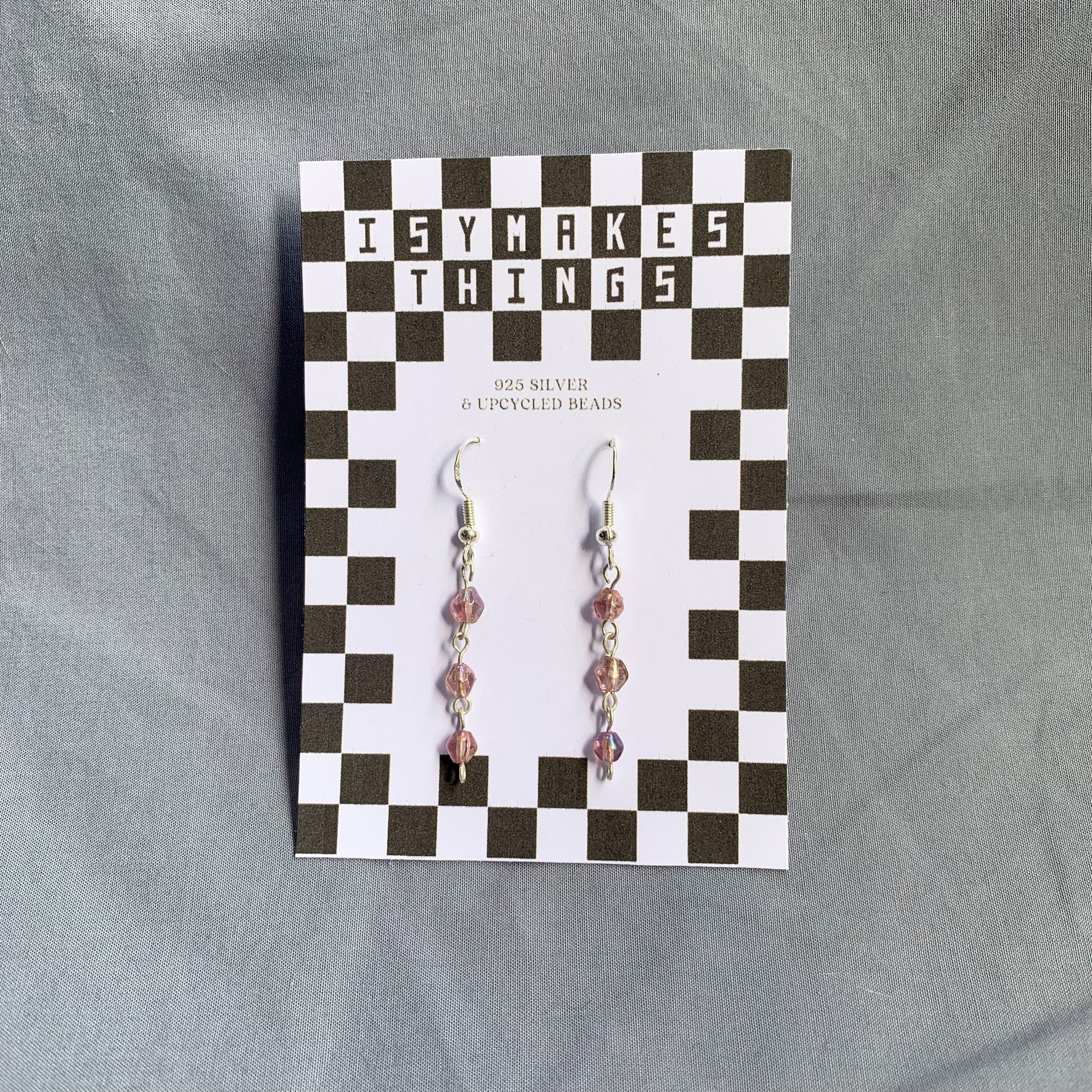 Three small purple gem like beads connected in a chain earrings on checkerboard backing card against a blue background