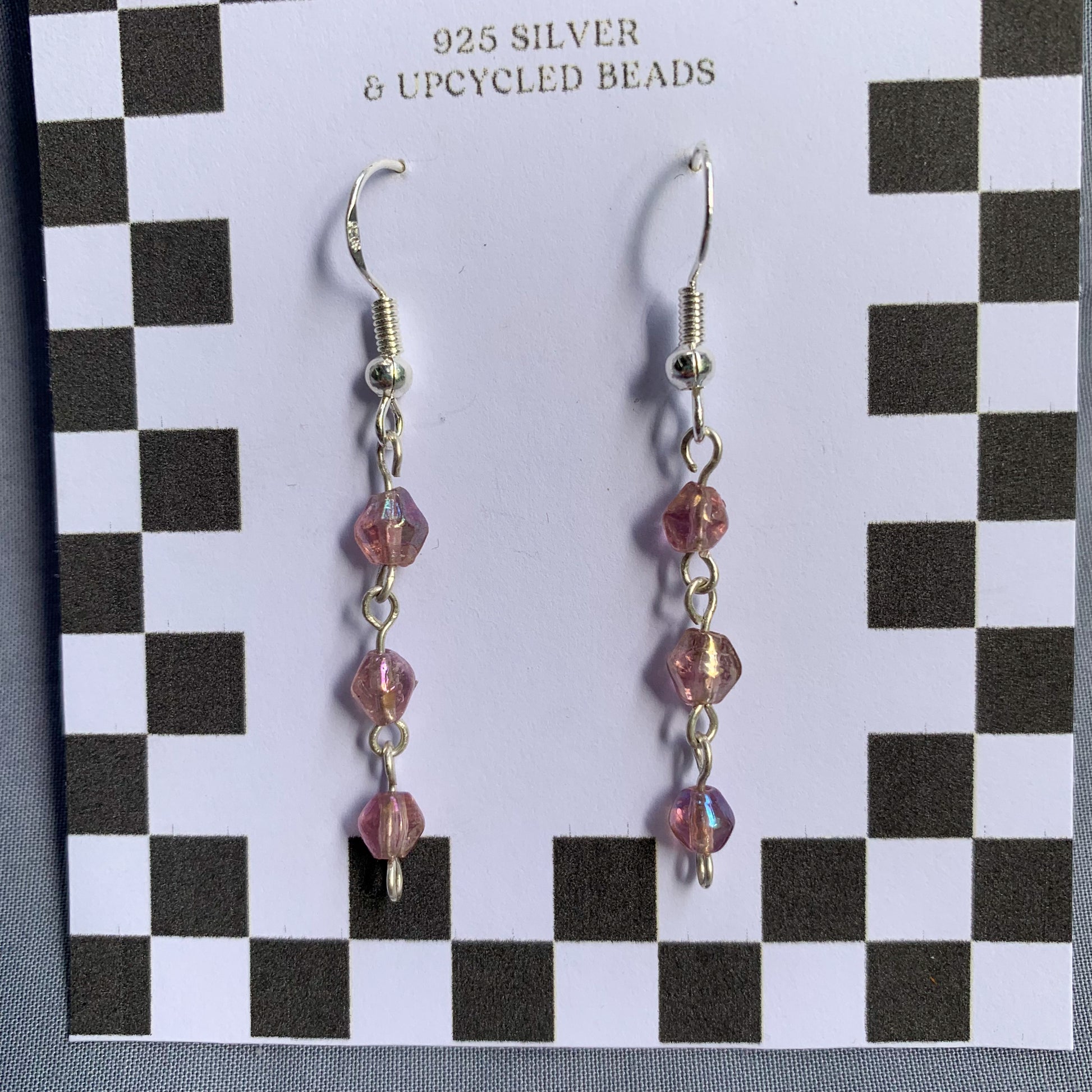 Three small purple gem like beads connected in a chain earrings on checkerboard backing card against a blue background