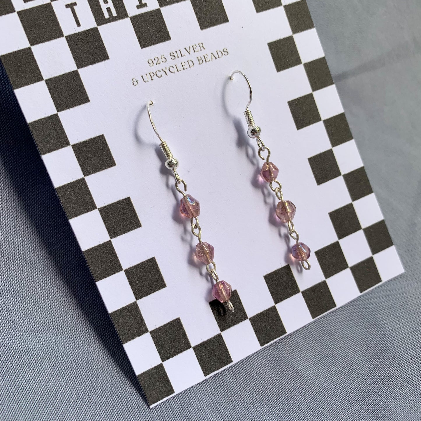 Three small purple gem like beads connected in a chain earrings on checkerboard backing card against a blue background
