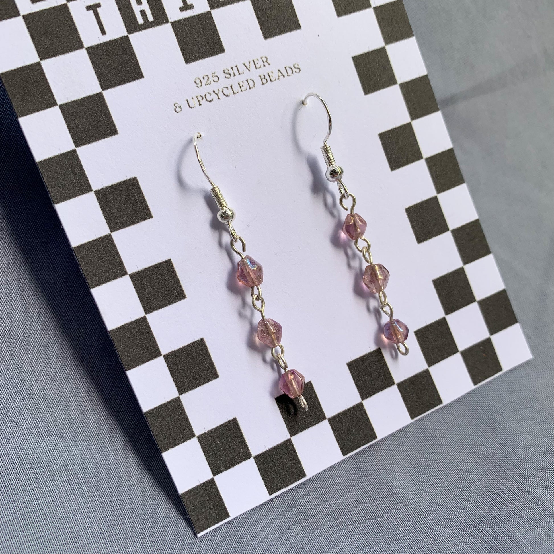 Three small purple gem like beads connected in a chain earrings on checkerboard backing card against a blue background