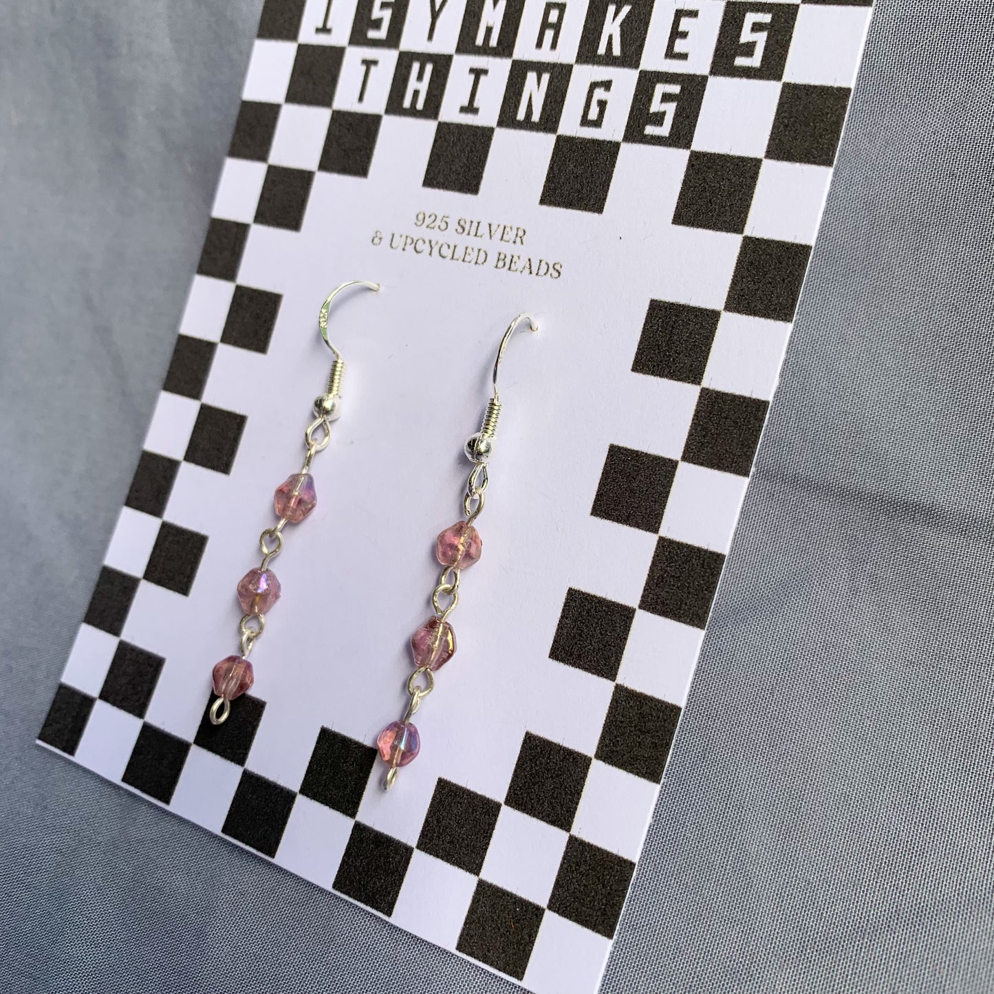 Three small purple gem like beads connected in a chain earrings on checkerboard backing card against a blue background