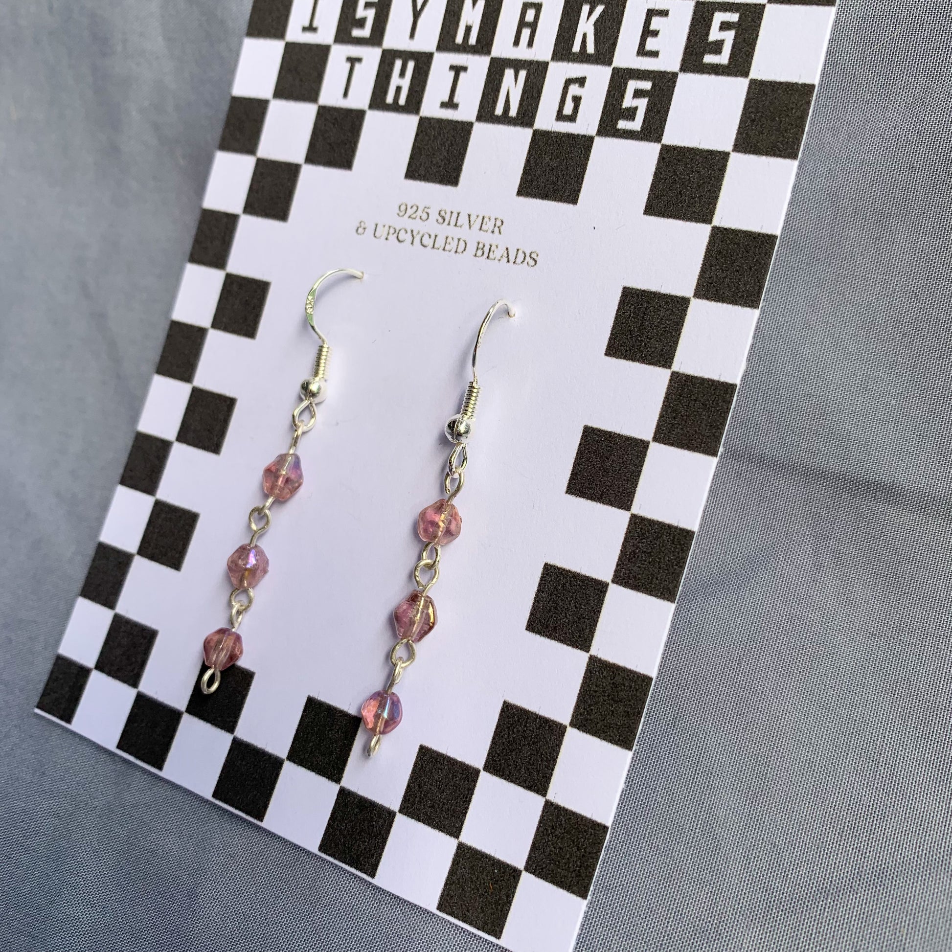 Three small purple gem like beads connected in a chain earrings on checkerboard backing card against a blue background