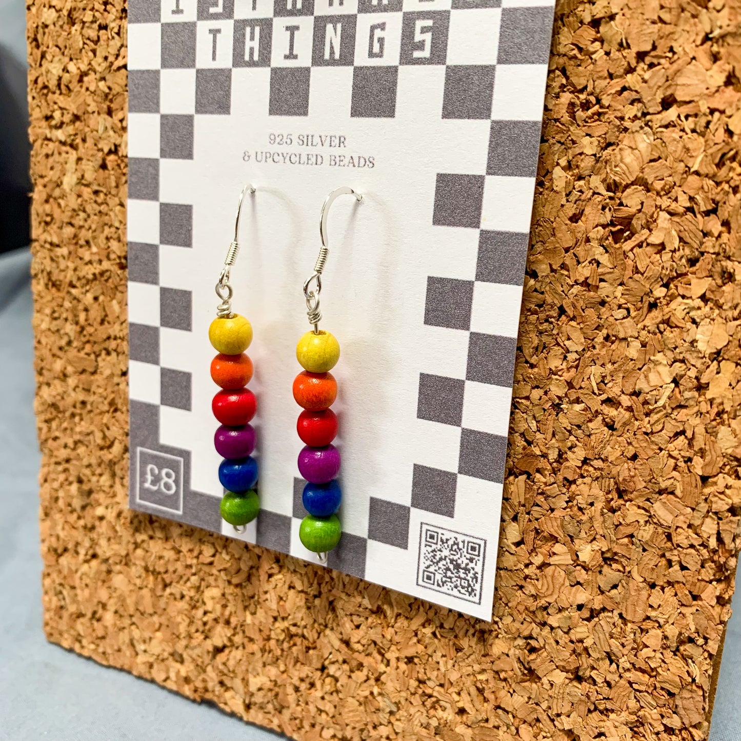 Rainbow bead earrings on a checkerboard backing pegged to a corkboard against a blue background