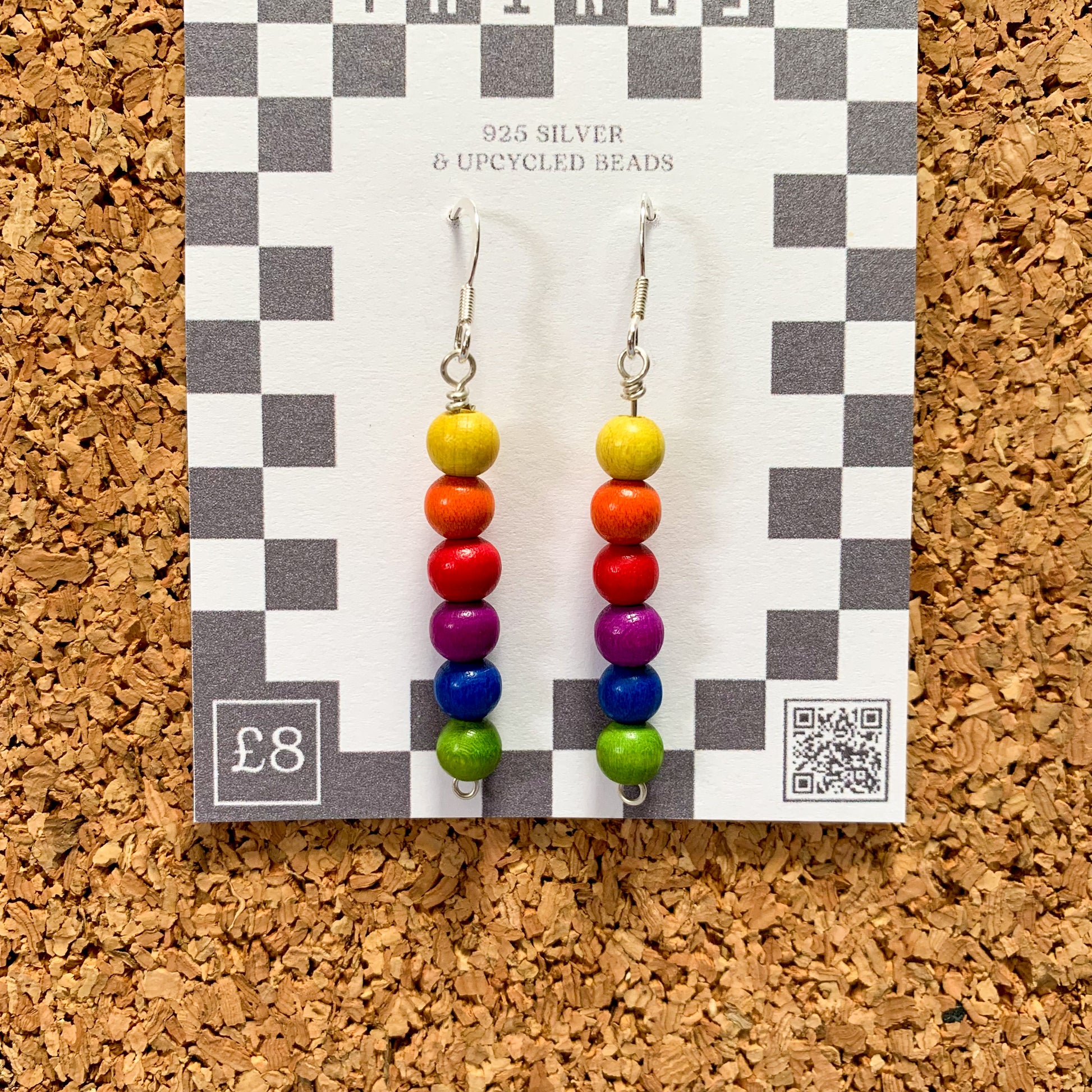 Rainbow bead earrings on a checkerboard backing pegged to a corkboard against a blue background