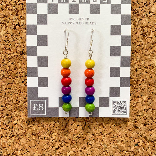 Rainbow bead earrings on a checkerboard backing pegged to a corkboard against a blue background