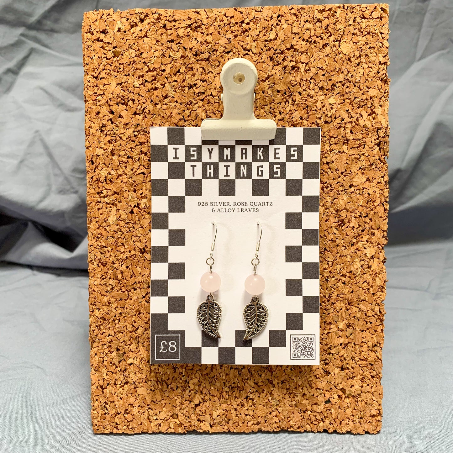 Rose quartz bead and leaf charm earrings on a checkerboard backing pegged to a corkboard against a blue background