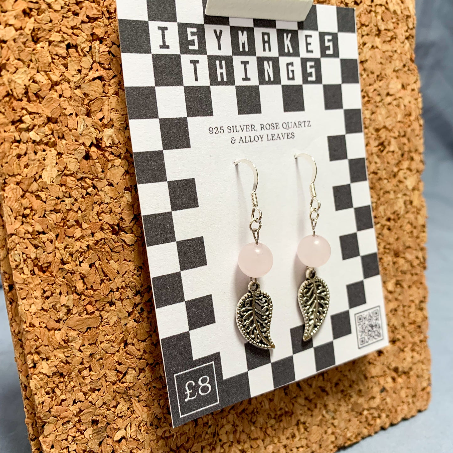 Rose quartz bead and leaf charm earrings on a checkerboard backing pegged to a corkboard against a blue background