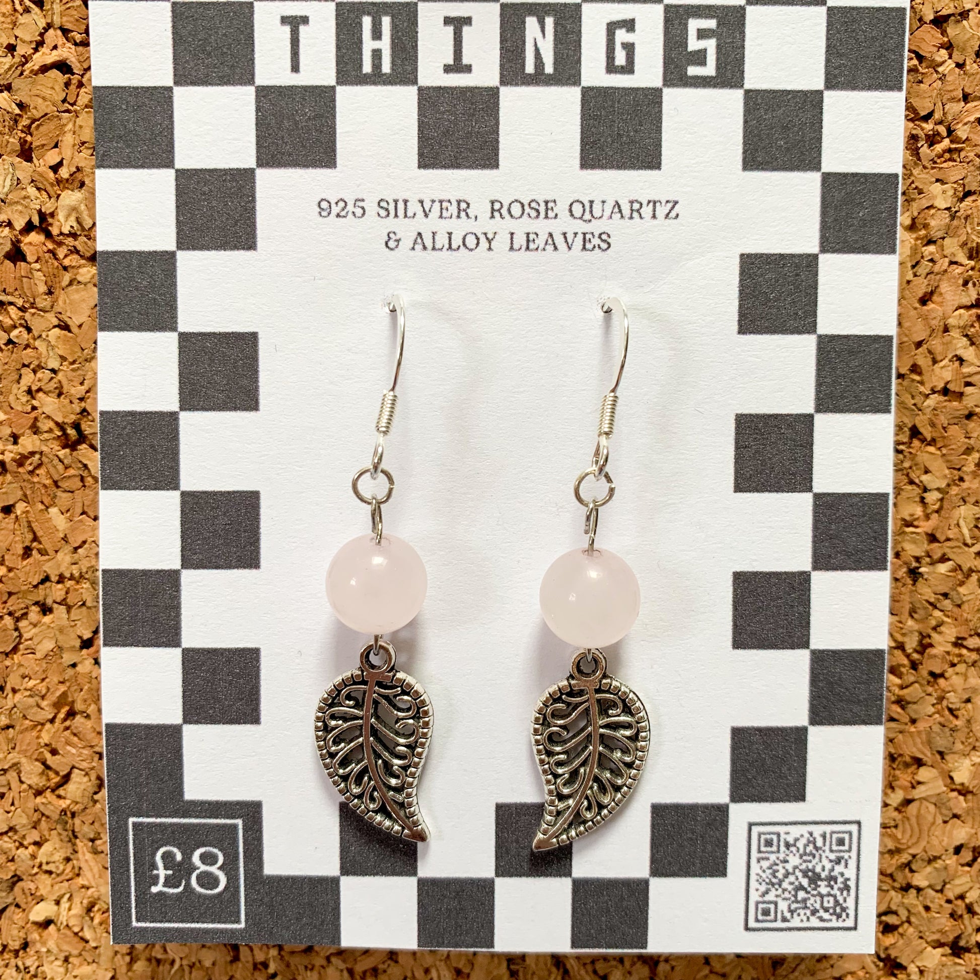 Rose quartz bead and leaf charm earrings on a checkerboard backing pegged to a corkboard against a blue background