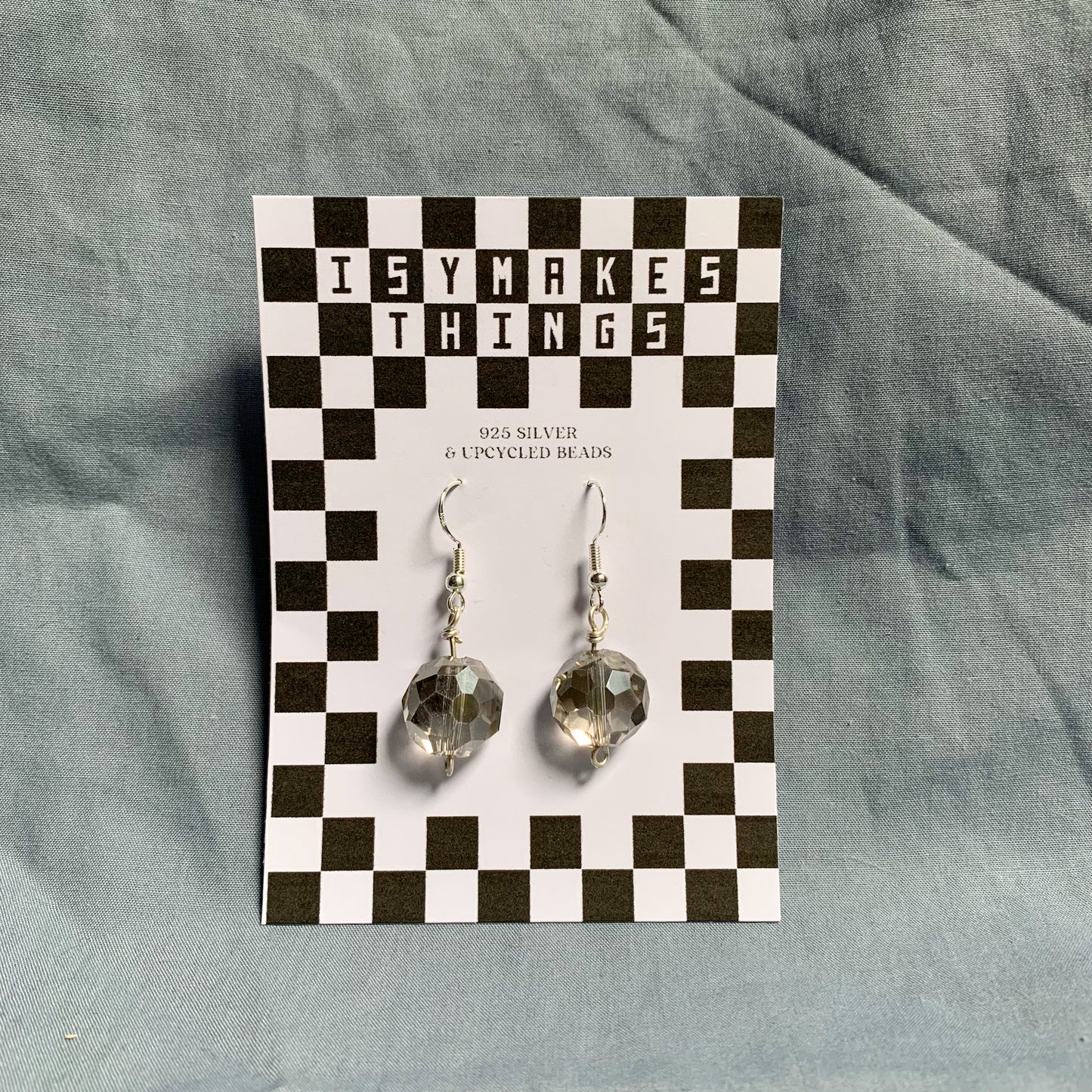 Shiny geometric clear ball earrings hang off a black and white checkered backing card with the title isymakesthings