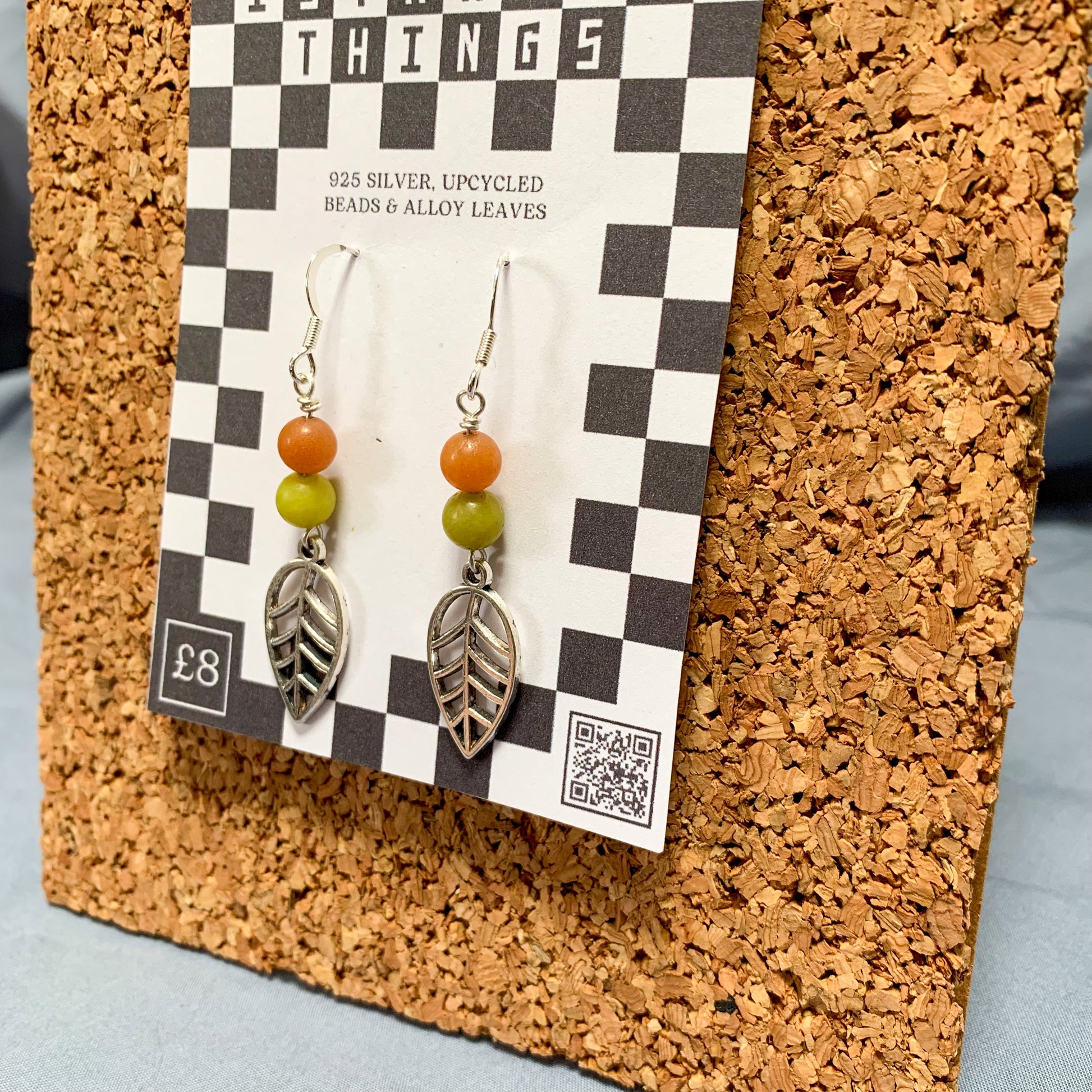 Orange & green leaf charm earrings on a checkerboard backing pegged to a corkboard against a blue background