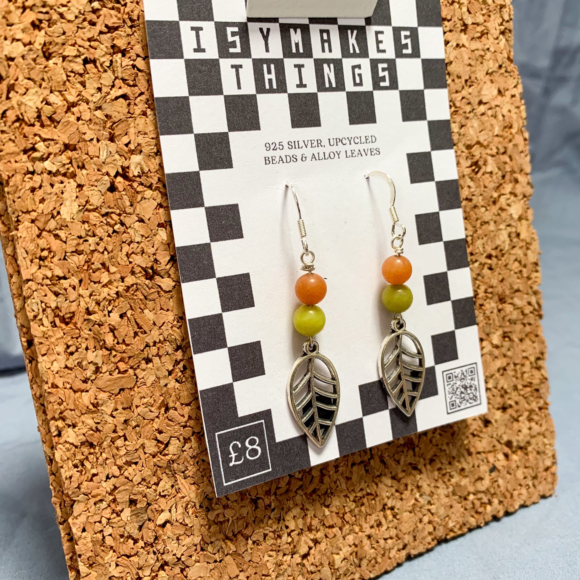 Orange & green leaf charm earrings on a checkerboard backing pegged to a corkboard against a blue background