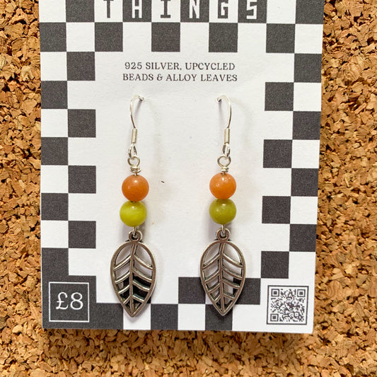 Orange & green leaf charm earrings on a checkerboard backing pegged to a corkboard against a blue background