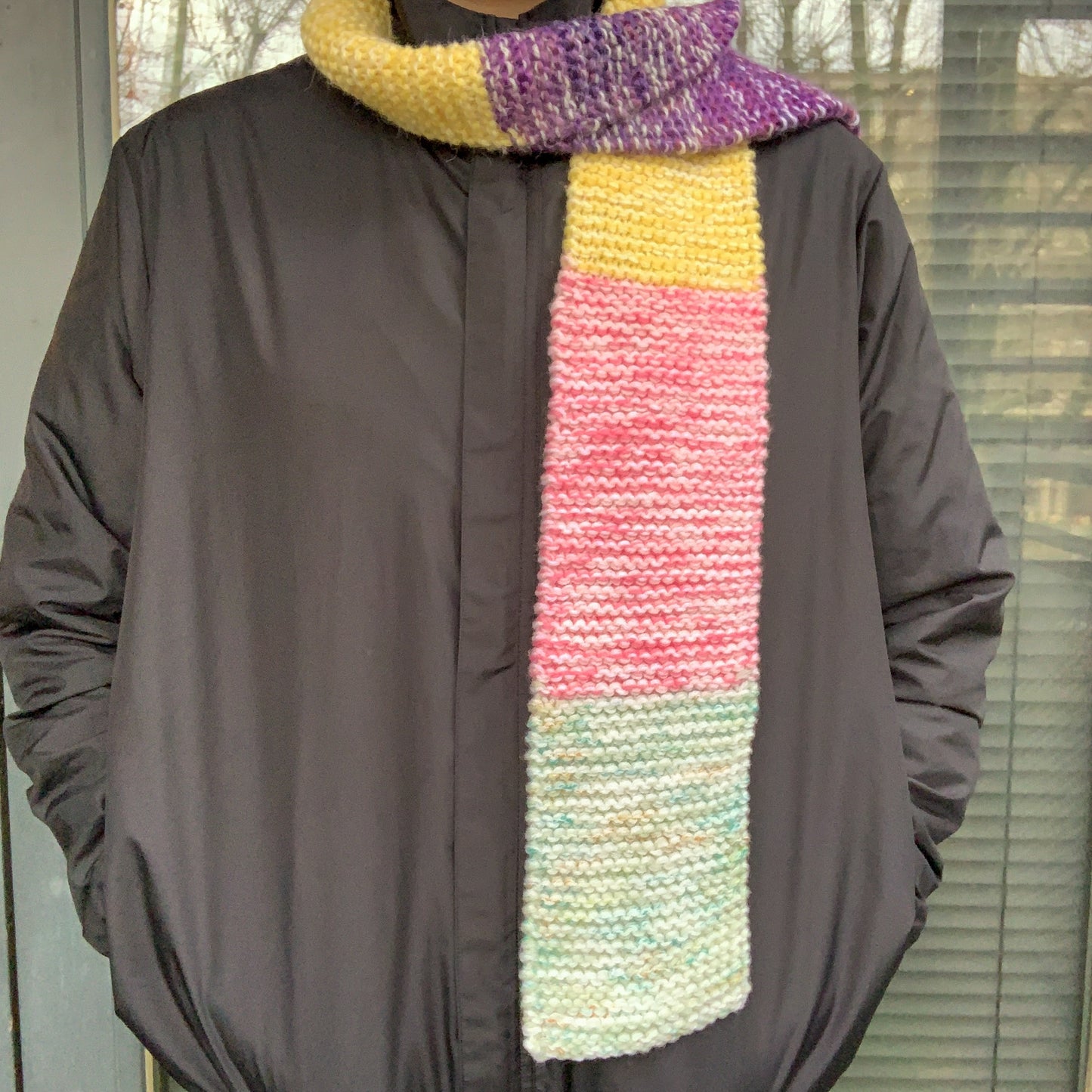 A green purple yellow and pink colour block knitted scarf is wrapped round a persons neck, one side down their front and the other behind them