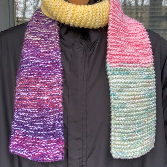 A green purple yellow and pink colour block knitted scarf is wrapped round a persons neck with it dangling down their front on both sides