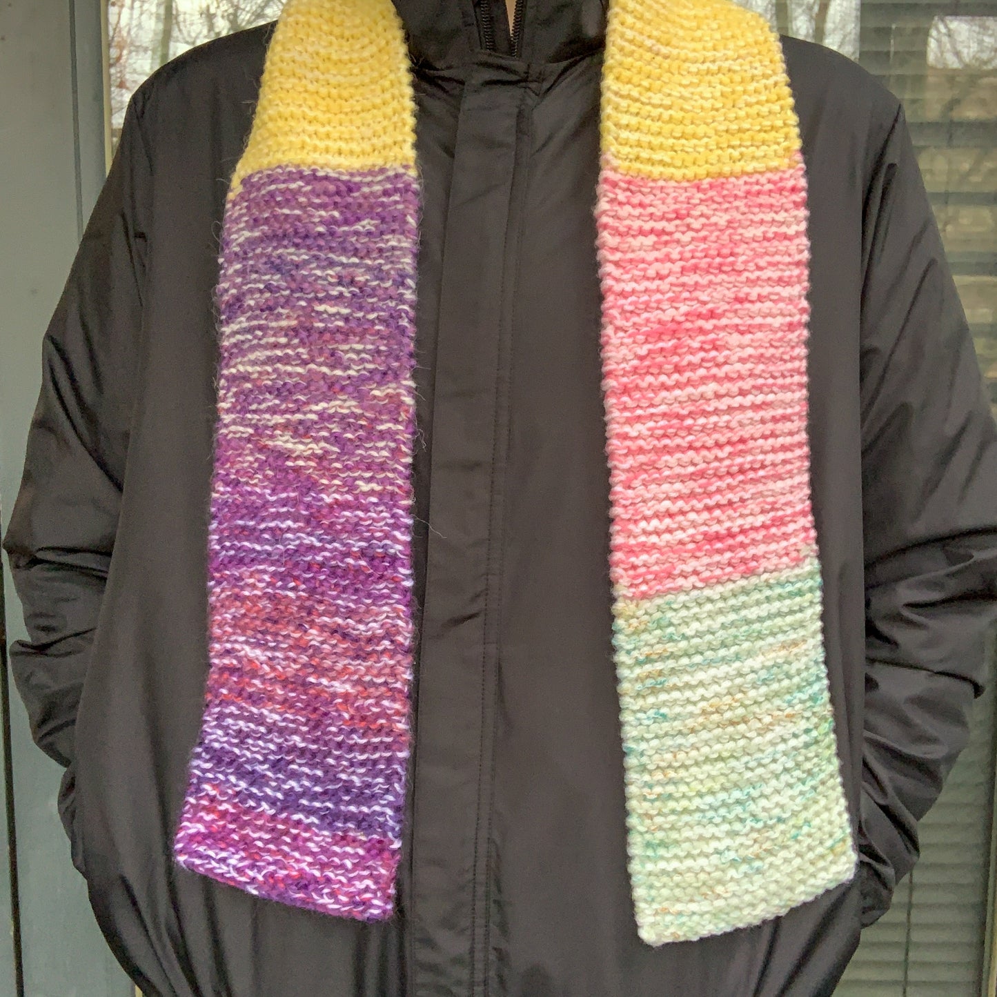 A green purple yellow and pink colour block knitted scarf is laid over a persons neck with it dangling down their front on both sides