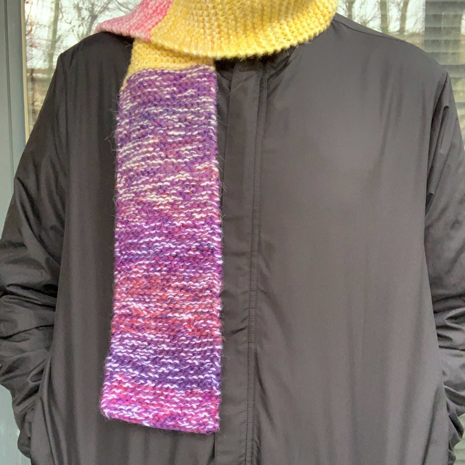 A green purple yellow and pink colour block knitted scarf is wrapped round a persons neck, one side down their front and the other behind them