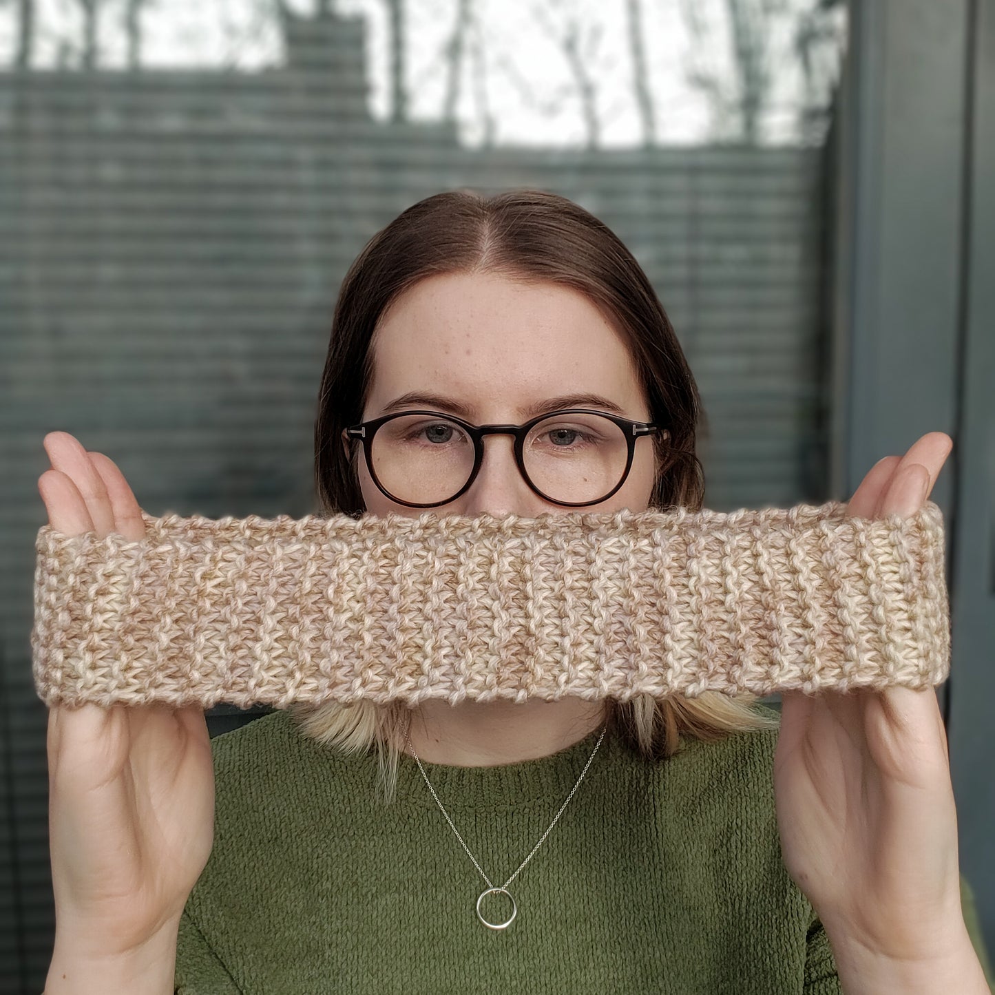 A woman holds a beige variegated ear warmer knitted headband stretched widely between her hands