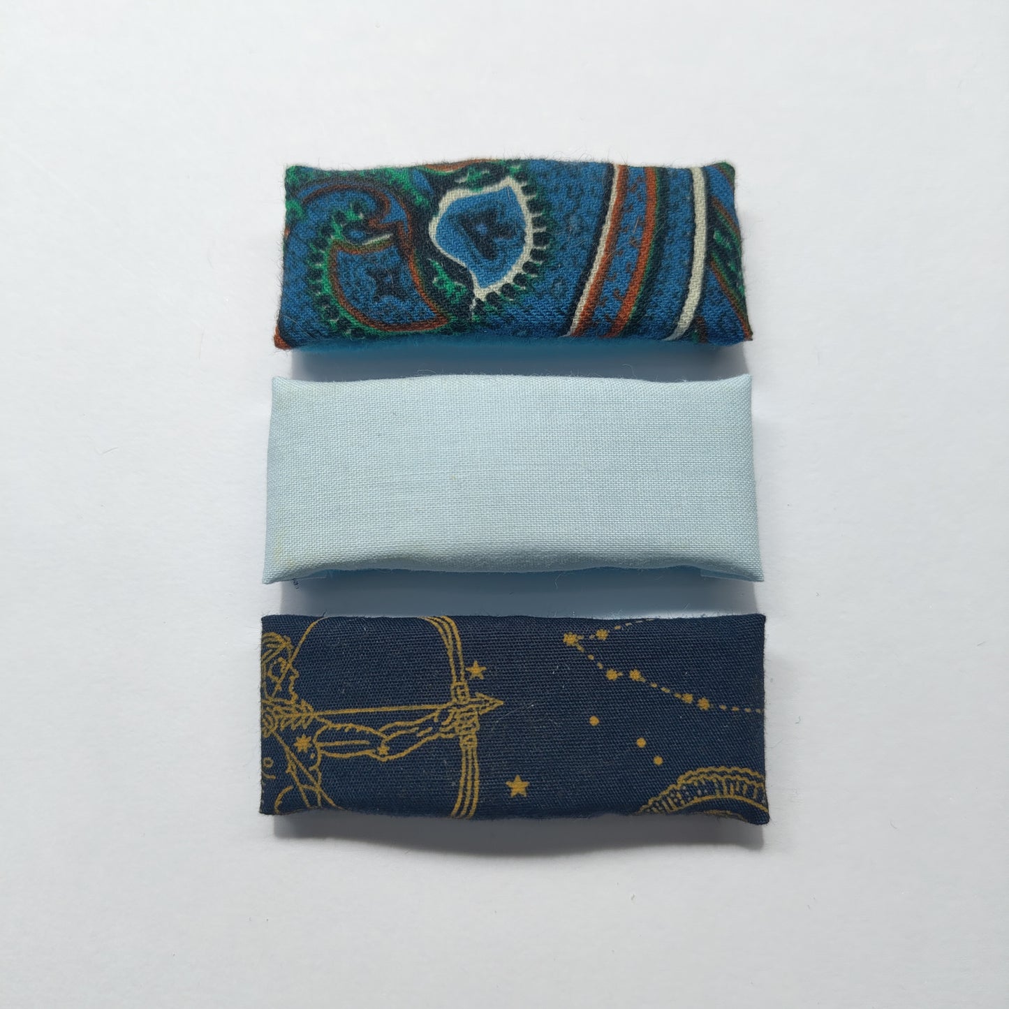 A trio of rectangular blue hair clips; floral paisley, plain and astrology, on a plain white background.