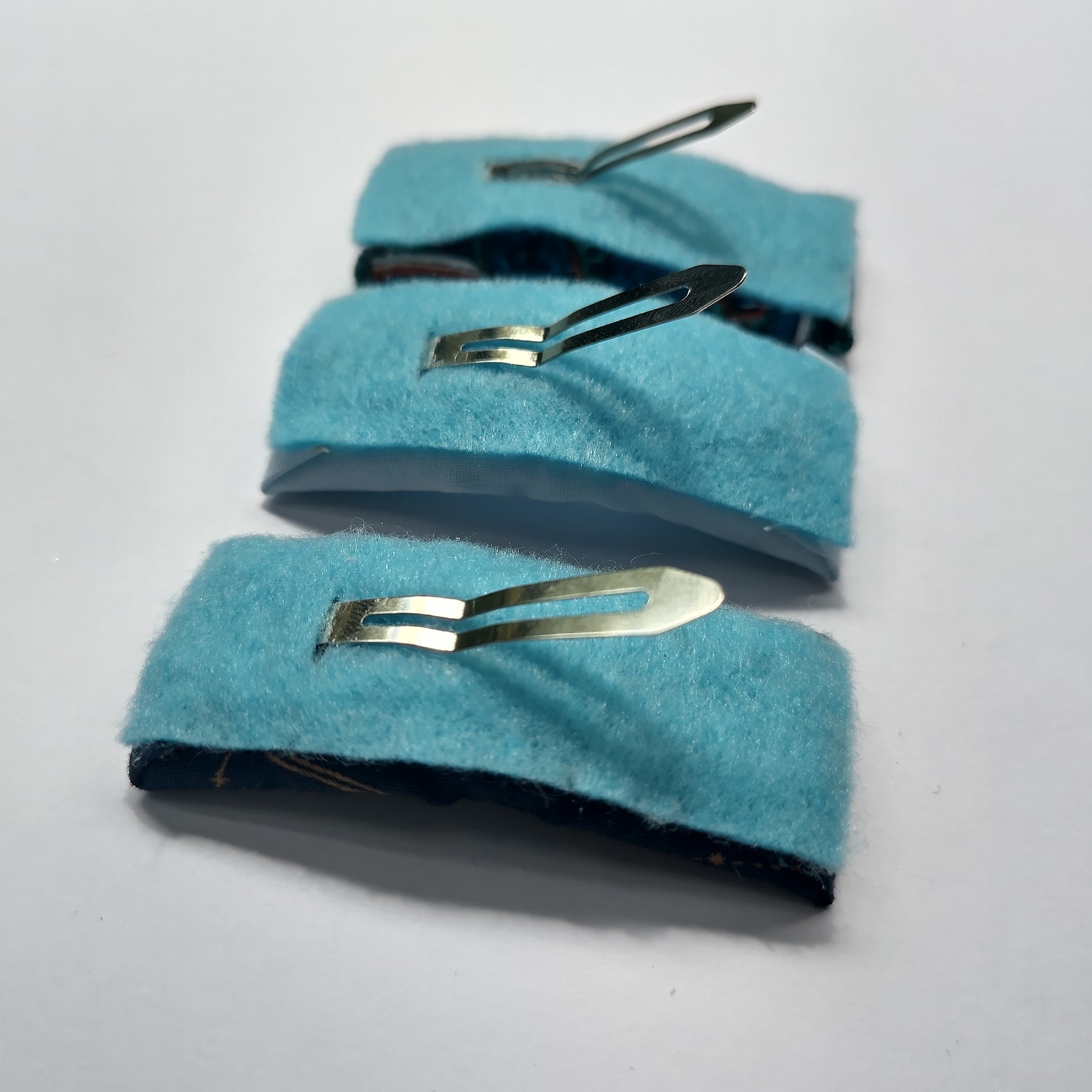 Three rectangular hair clips upside down displaying blue felt underneath and a silver prong.