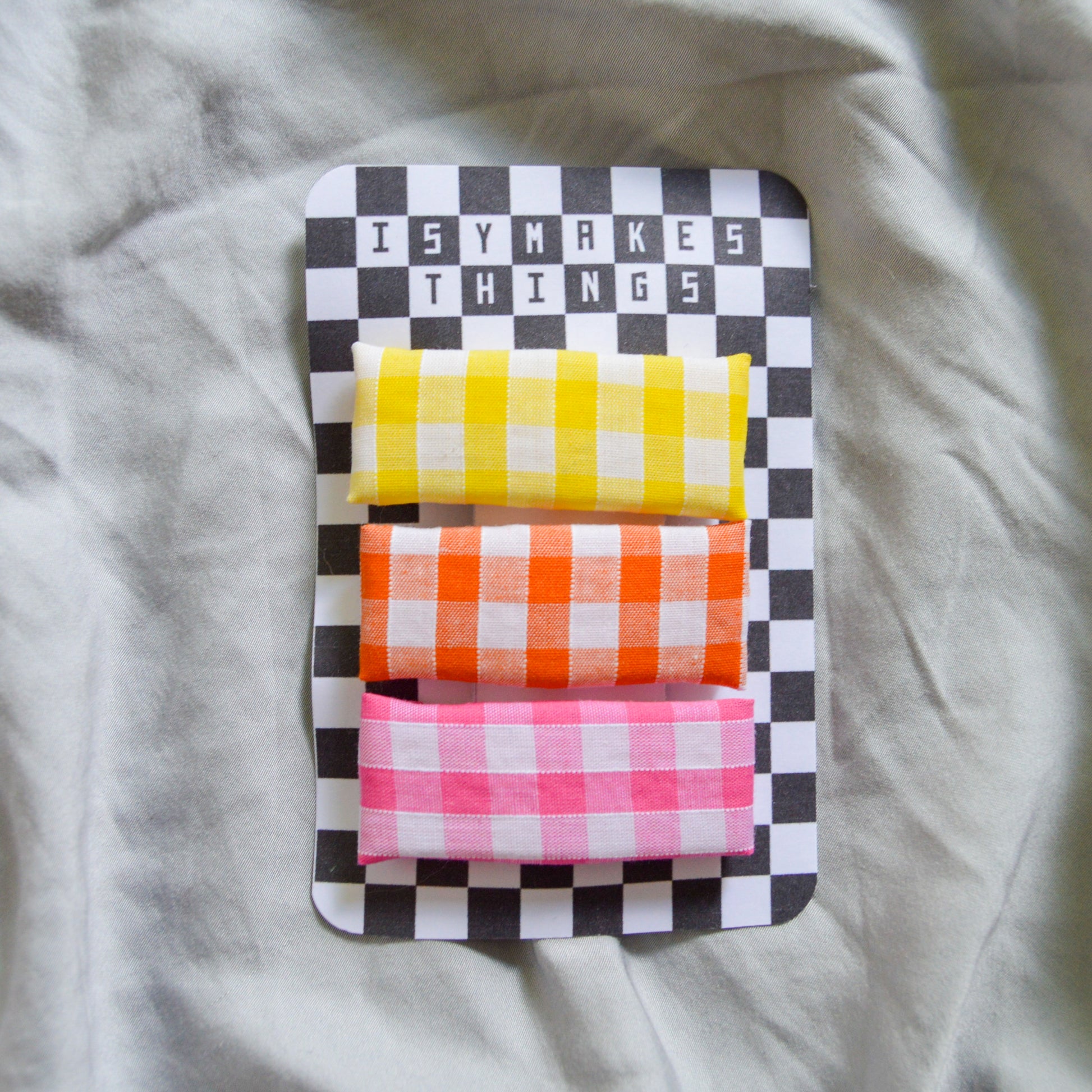 a trio of rectangle gingham hair clips, yellow orange and pink are clipped to a black and white checkerboard backing card with the title isymakesthings