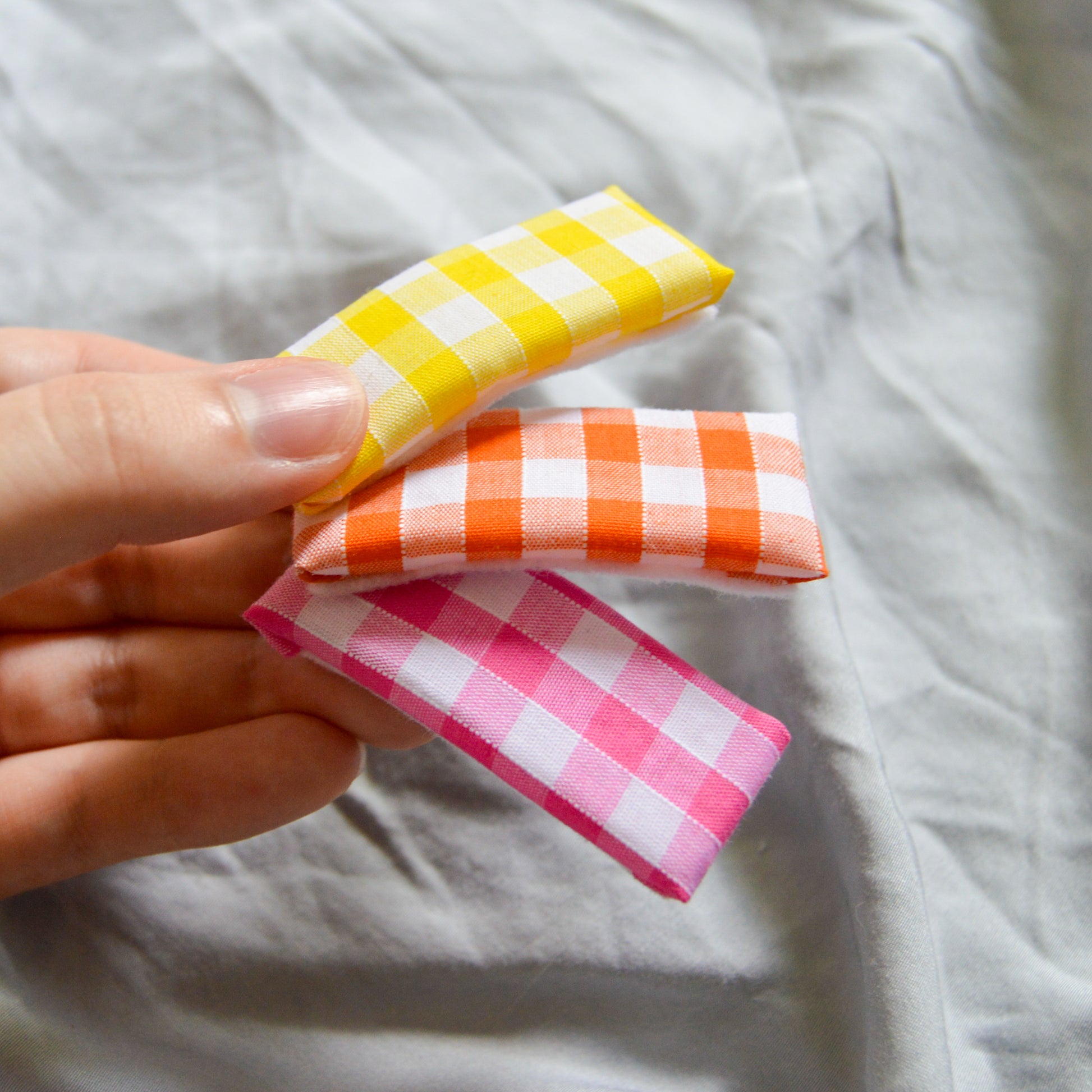 a trio of rectangle gingham hair clips, yellow orange and pink are fanned out and held between a thumb and finger