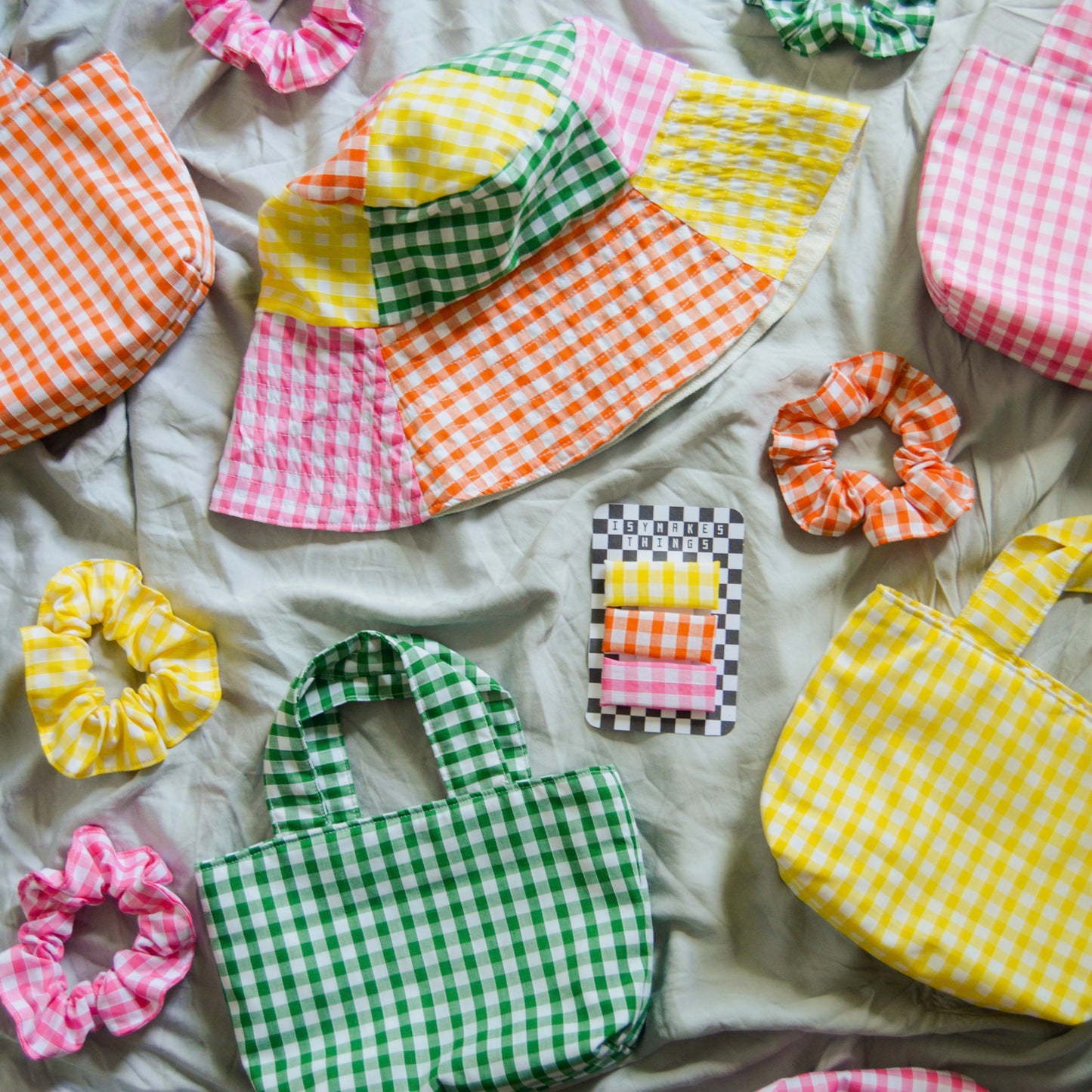 a collection of green yellow orange and pink gingham accessories are arranged together, including a multicolour patchwork sun hat, mini tote bags, scrunchies and hair clips