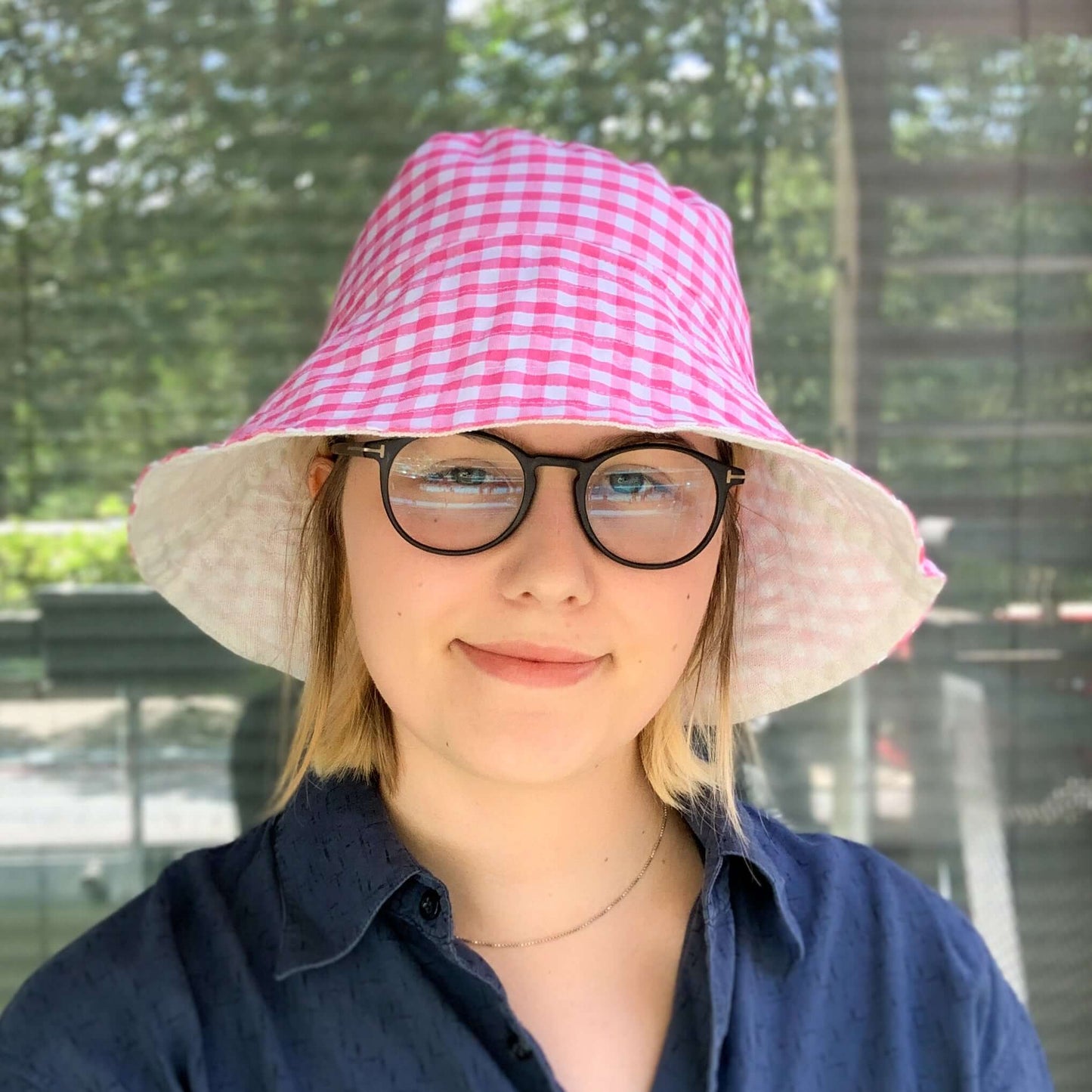 A woman wearing a dark blue shirt and glasses models a bright pink gingham bucket sun hat while smiling