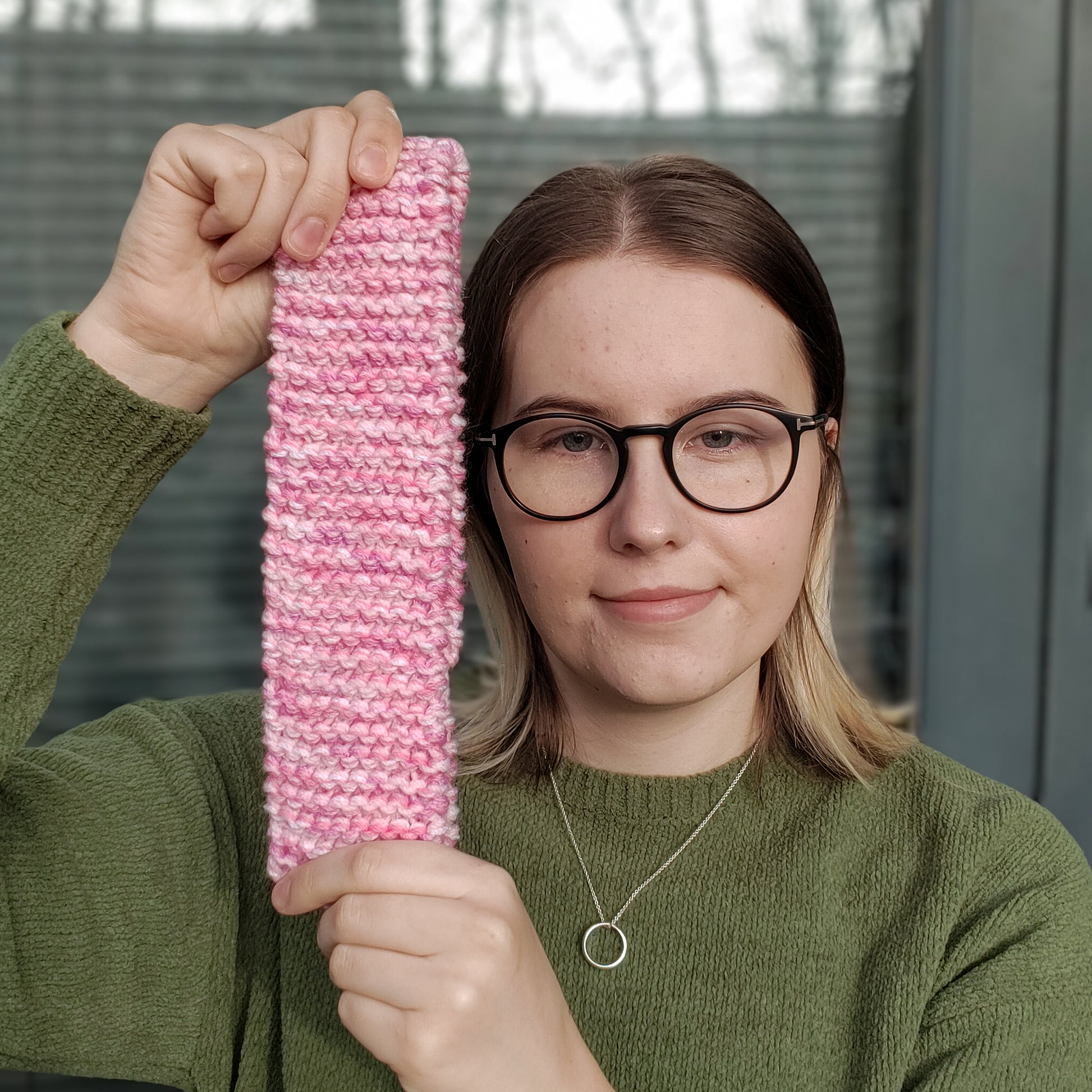 A woman in a green jumper and glasses holds a pink variegated knitted ear warmer headband while smiling