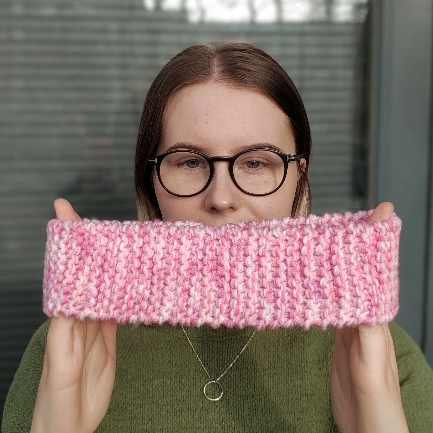 A woman in a green jumper and glasses holds a pink variegated knitted ear warmer headband between her hands in front of herself