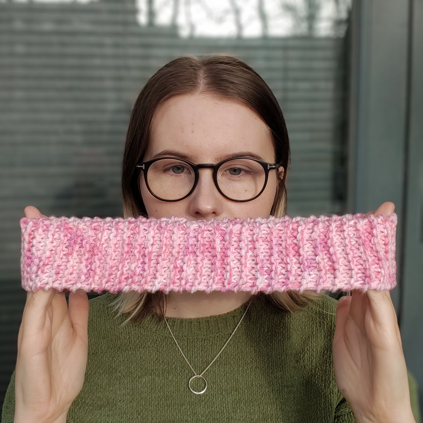 A woman in a green jumper and glasses holds a pink variegated knitted ear warmer headband stretched out between her hands in front of herself