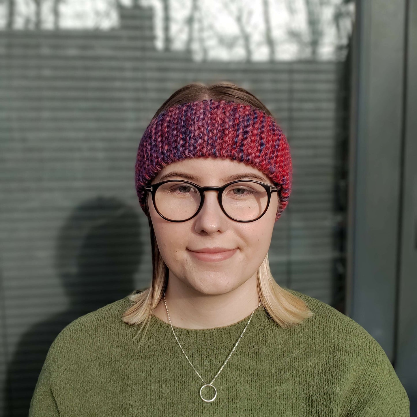 A woman wearing a green jumper and glasses models a red and purple variegated gradient knitted headband ear warmer