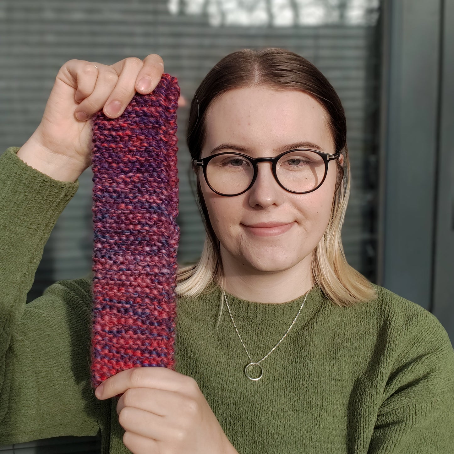 A woman wearing a green jumper and glasses holds out a red and purple variegated gradient knitted headband ear warmer