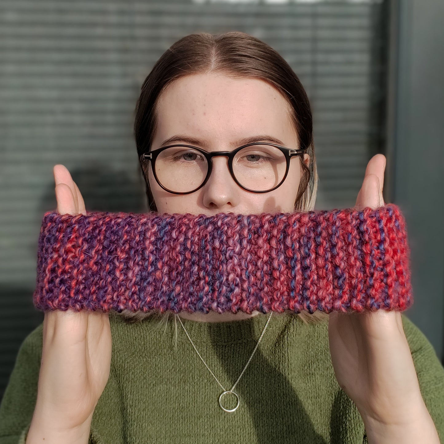 A woman wearing a green jumper and glasses holds a red and purple variegated gradient knitted headband ear warmer between her hands