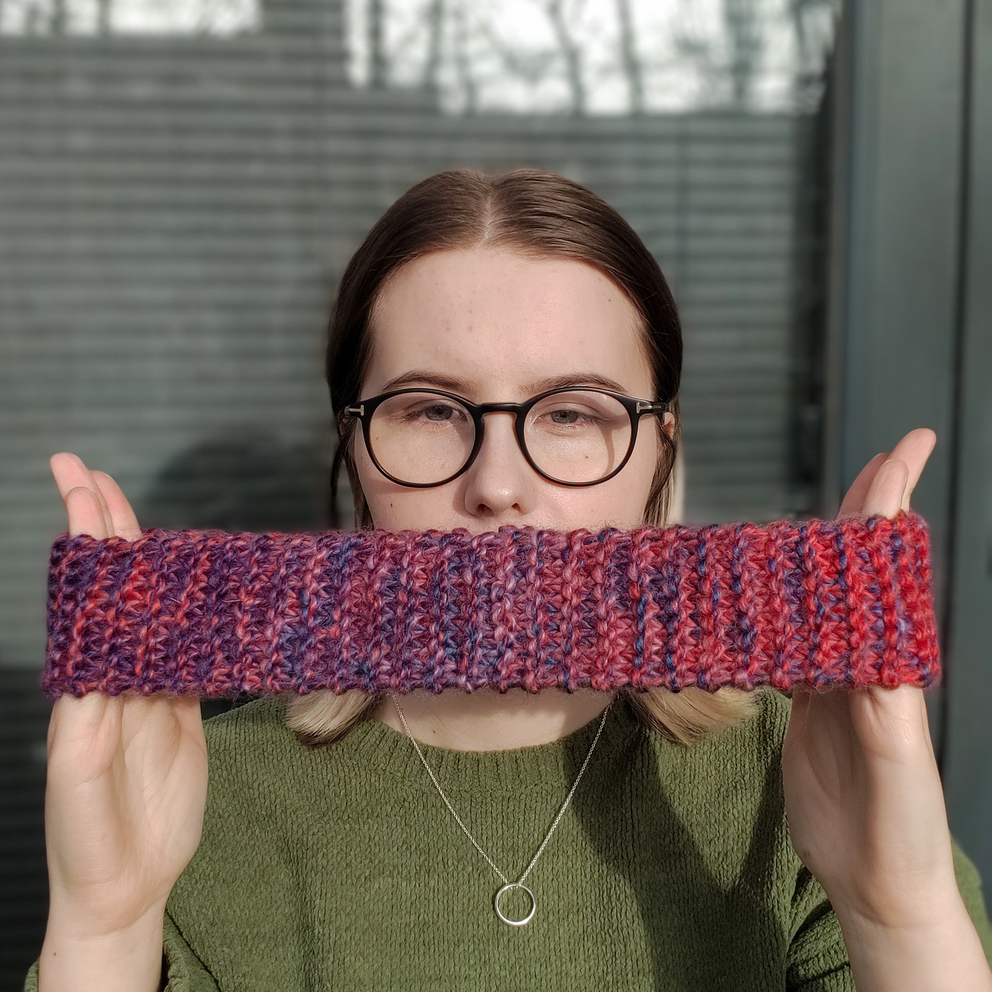 A woman wearing a green jumper and glasses holds a red and purple variegated gradient knitted headband ear warmer stretched out between her hands in front of her