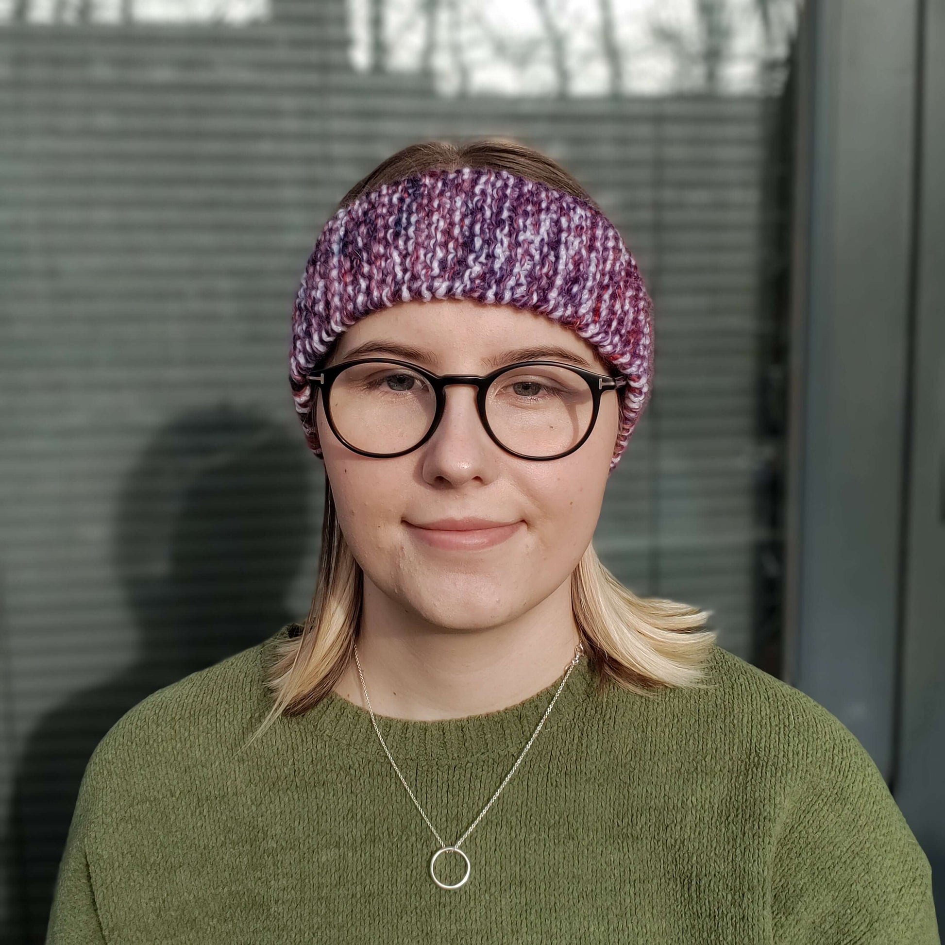 A woman in a green jumper and glasses models a white purple & red variegated knitted headband ear warmer