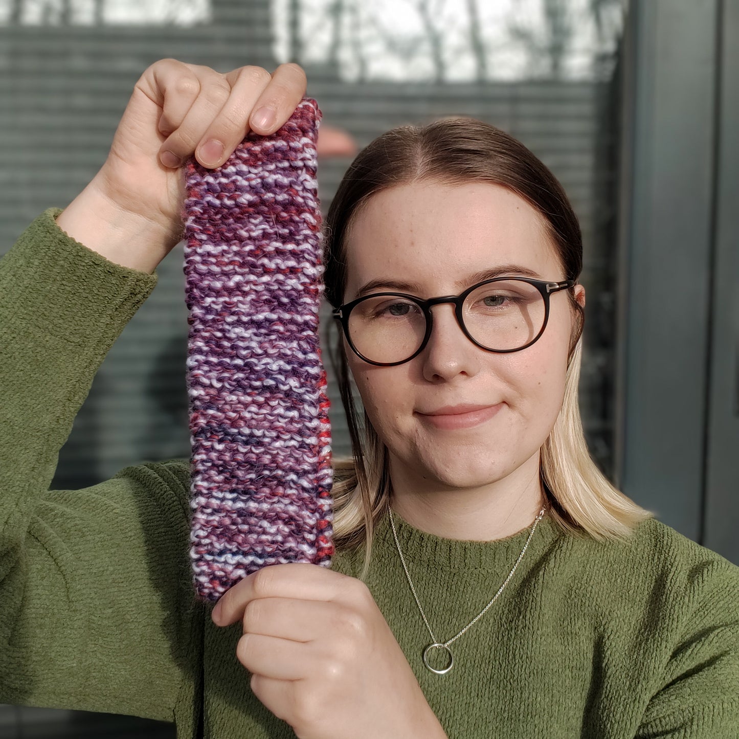 A woman in a green jumper and glasses holds up a white purple & red variegated knitted headband ear warmer