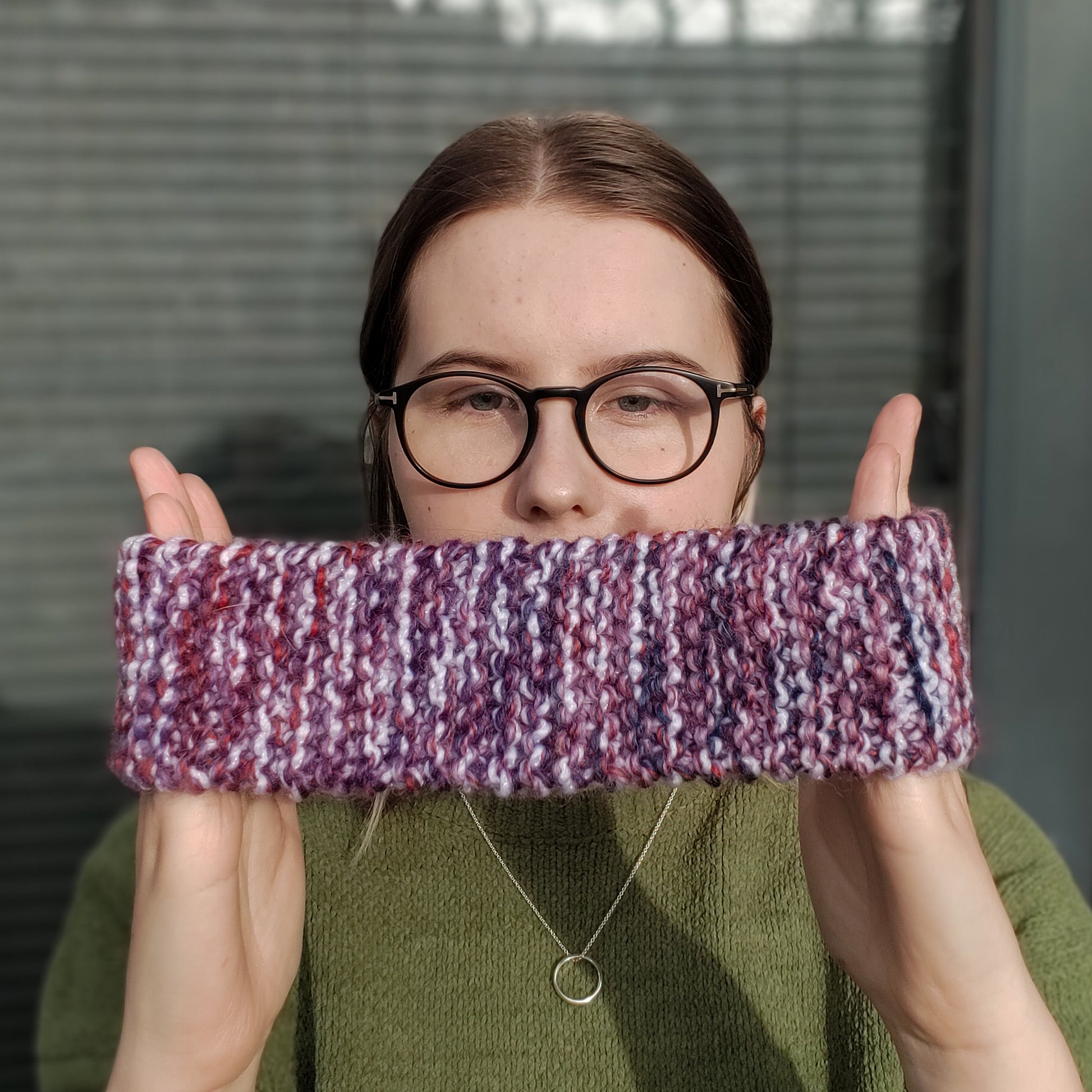 A woman in a green jumper and glasses holds a white purple & red variegated knitted headband ear warmer between her hands