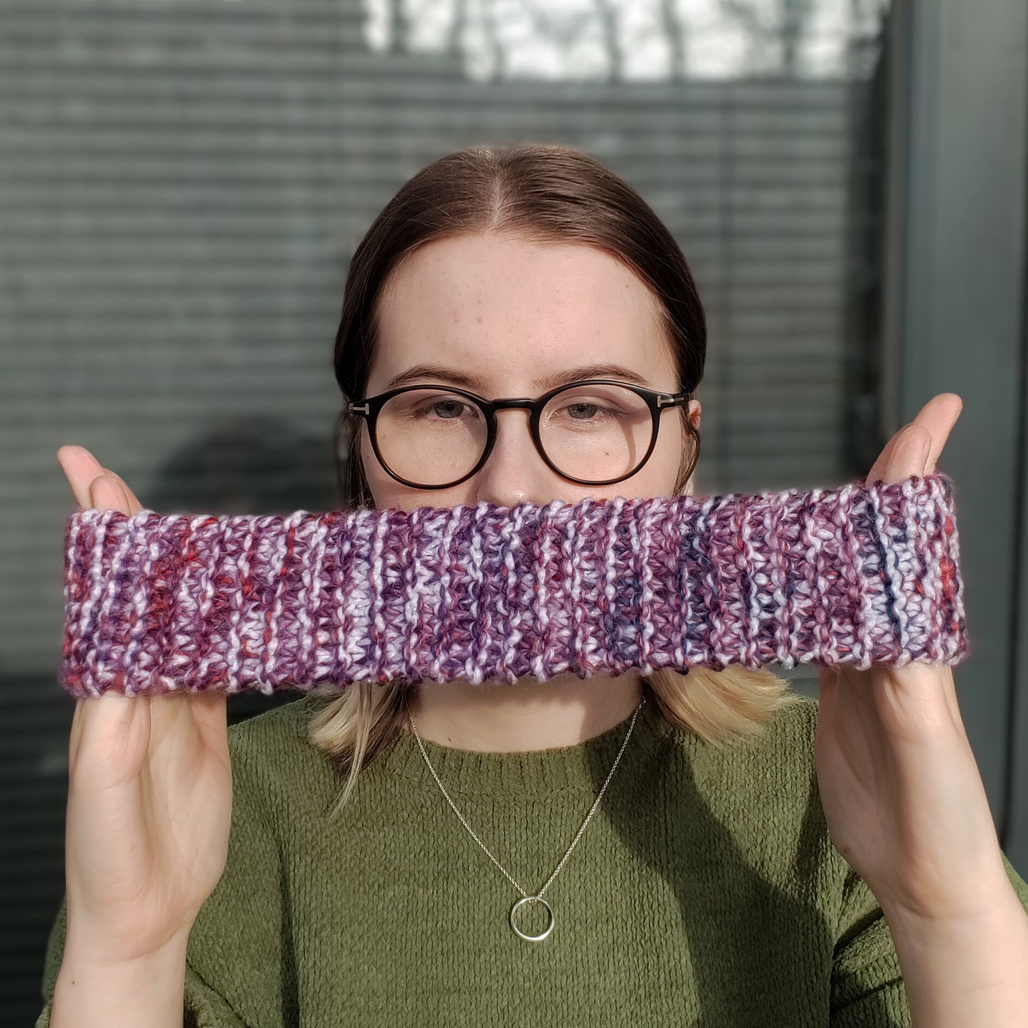 A woman in a green jumper and glasses holds a white purple & red variegated knitted headband ear warmer stretched between her hands