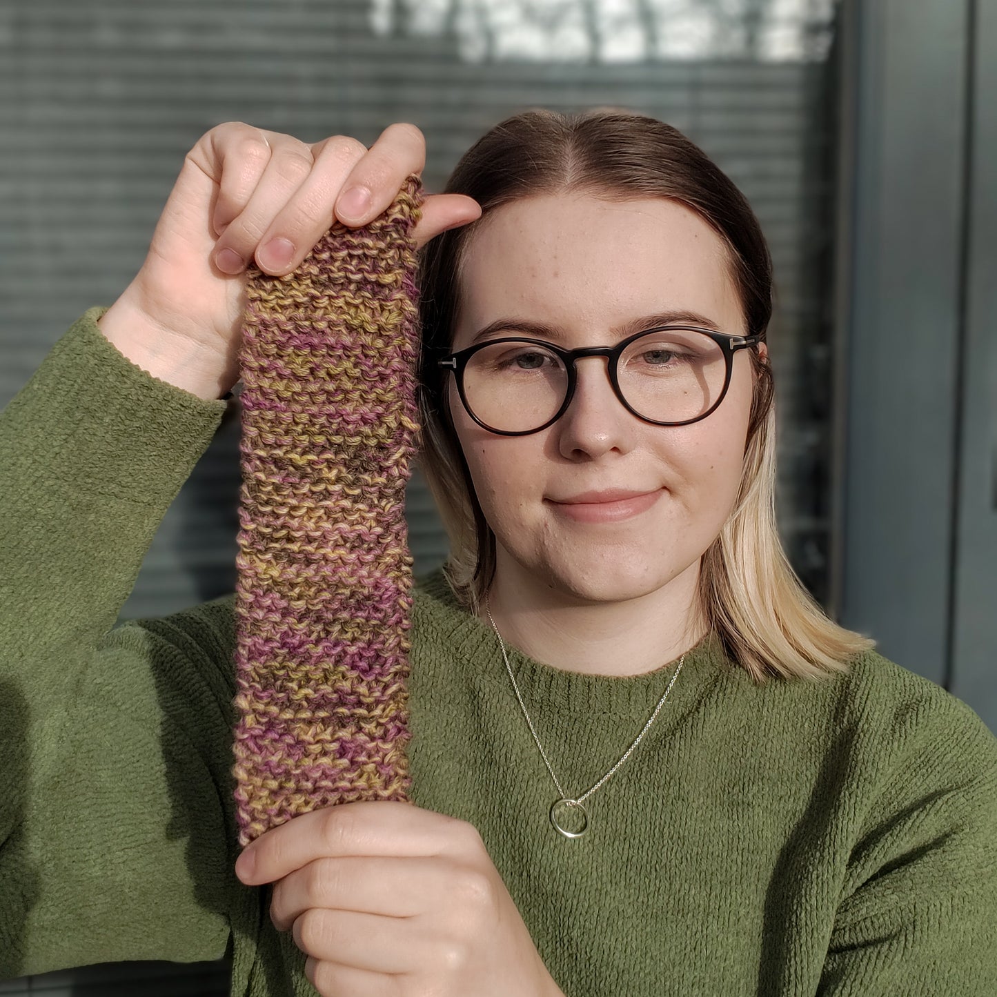 A blonde woman in a green jumper and glasses holds up a dark yellow green and purple variegated knitted headband 