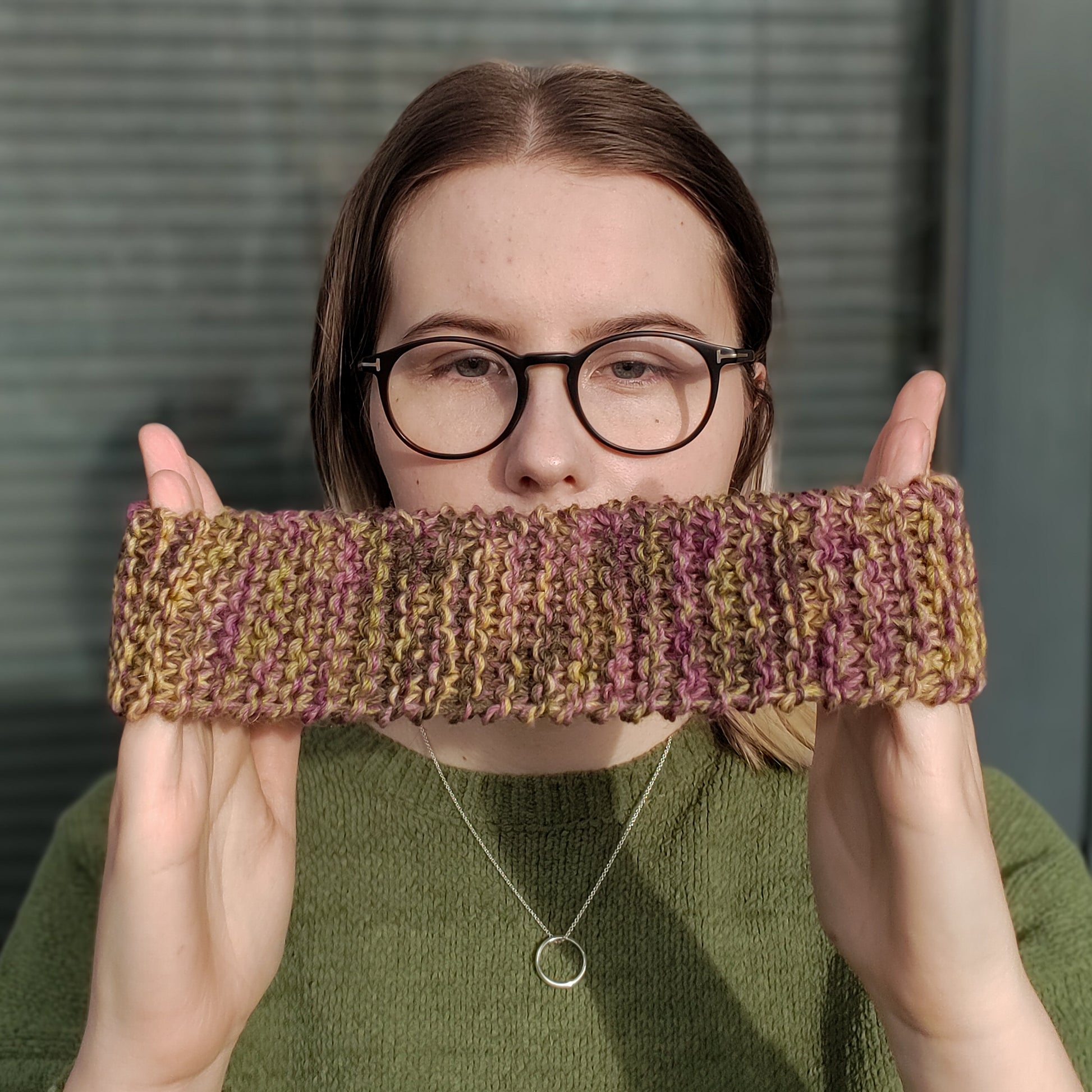 A blonde woman in a green jumper and glasses holds a dark yellow green and purple variegated knitted headband between her hands