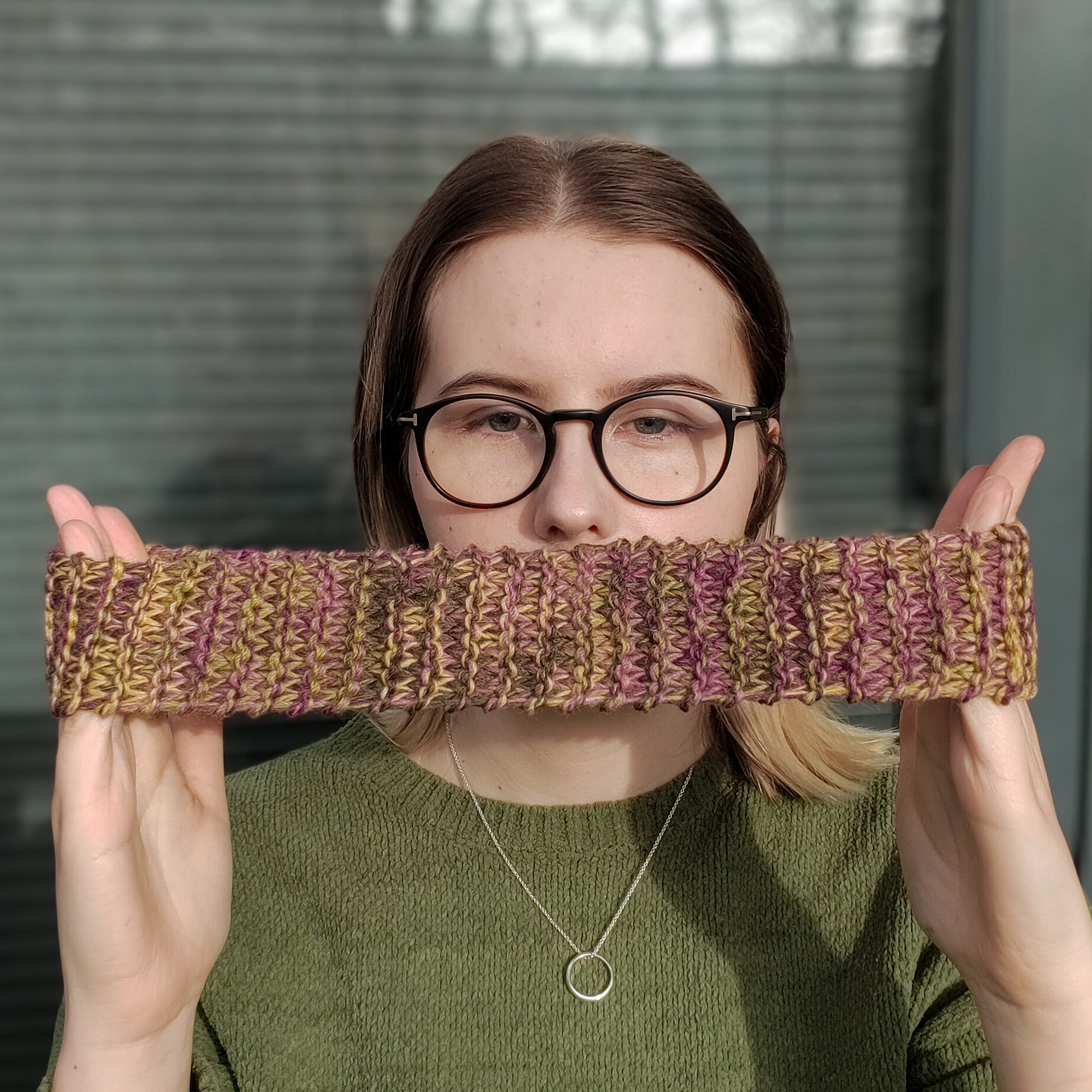 A blonde woman in a green jumper and glasses holds a dark yellow green and purple variegated knitted headband stretched between her hands